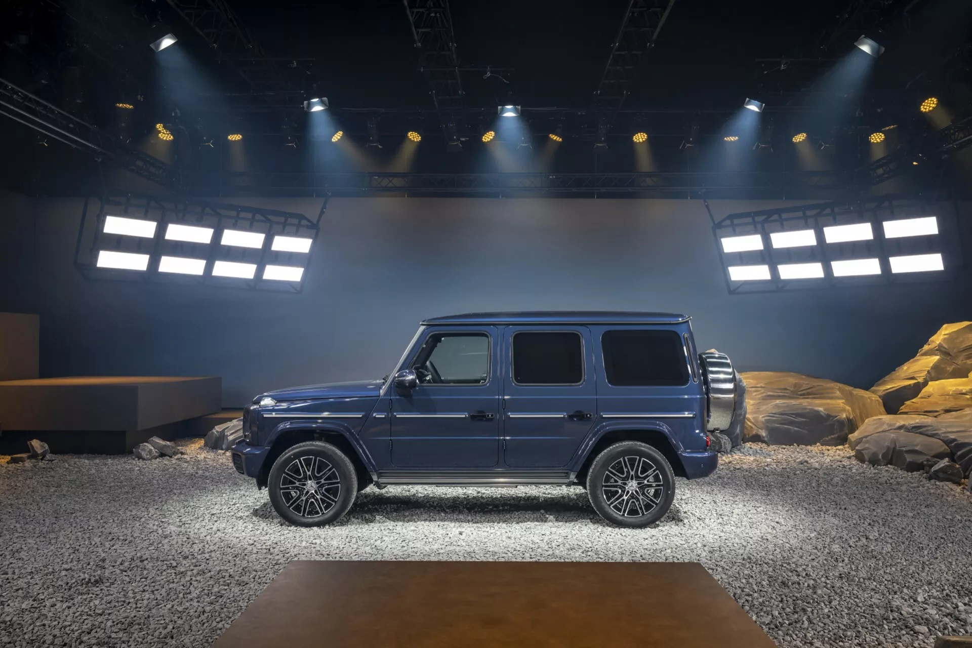 Bočný profil luxusného SUV Mercedes-Benz G-Klasse 465 ročník 2024 na simulovanom off-road vnútornom pódiu, zdôrazňujúci estetiku dizajnu, stabilitu a pokročilé technologické funkcie pod jasným osvetlením reflektorov