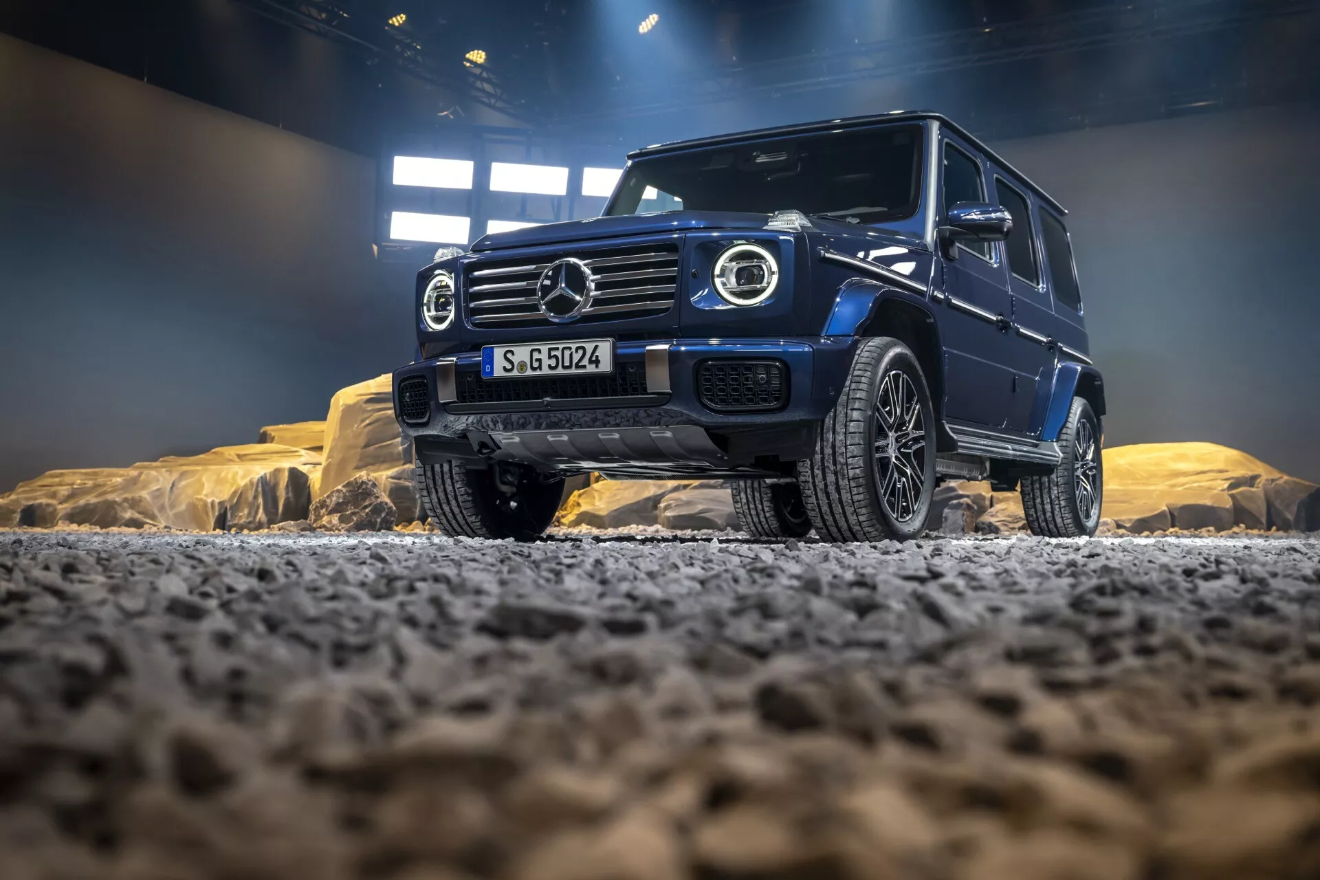 Mercedes-Benz triedy G (G 465) pre rok 2024 v terénnom prostredí, zdôrazňujúci jeho robustný postoj a luxusné prvky s výraznou mriežkou chladiča, LED svetlometmi a pokročilým dizajnom diskov kolies