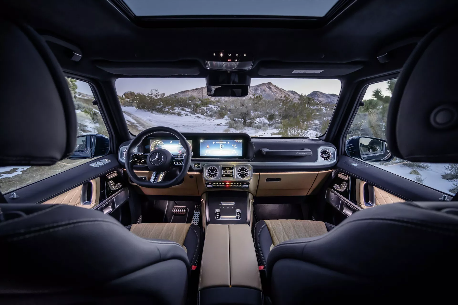 Pohľad na interiér Mercedes-Benz G-Class 2024 z zadného stredného miesta, s multifunkčným koženým volantom, duálnymi digitálnymi displejmi na prístrojovej doske, stredovou konzolou s dotykovým panelom, automatom na zmeny prevodov, čiernymi koženými sedadlami s elektronickým nastavením, prvky z dreva, okolitým osvetlením a čírym výhľadom cez predné čelné sklo