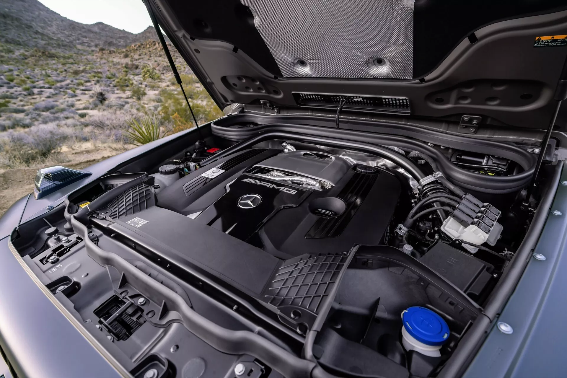 Detailný pohľad na motorový priestor Mercedes-Benz G-Klasse 2024 so znakom značky na kryte motora, detailné zobrazenie rôznych častí motora vrátane prívodu vzduchu a zásobníka na chladiacu kvapalinu, na pozadí s vonkajším prostredím