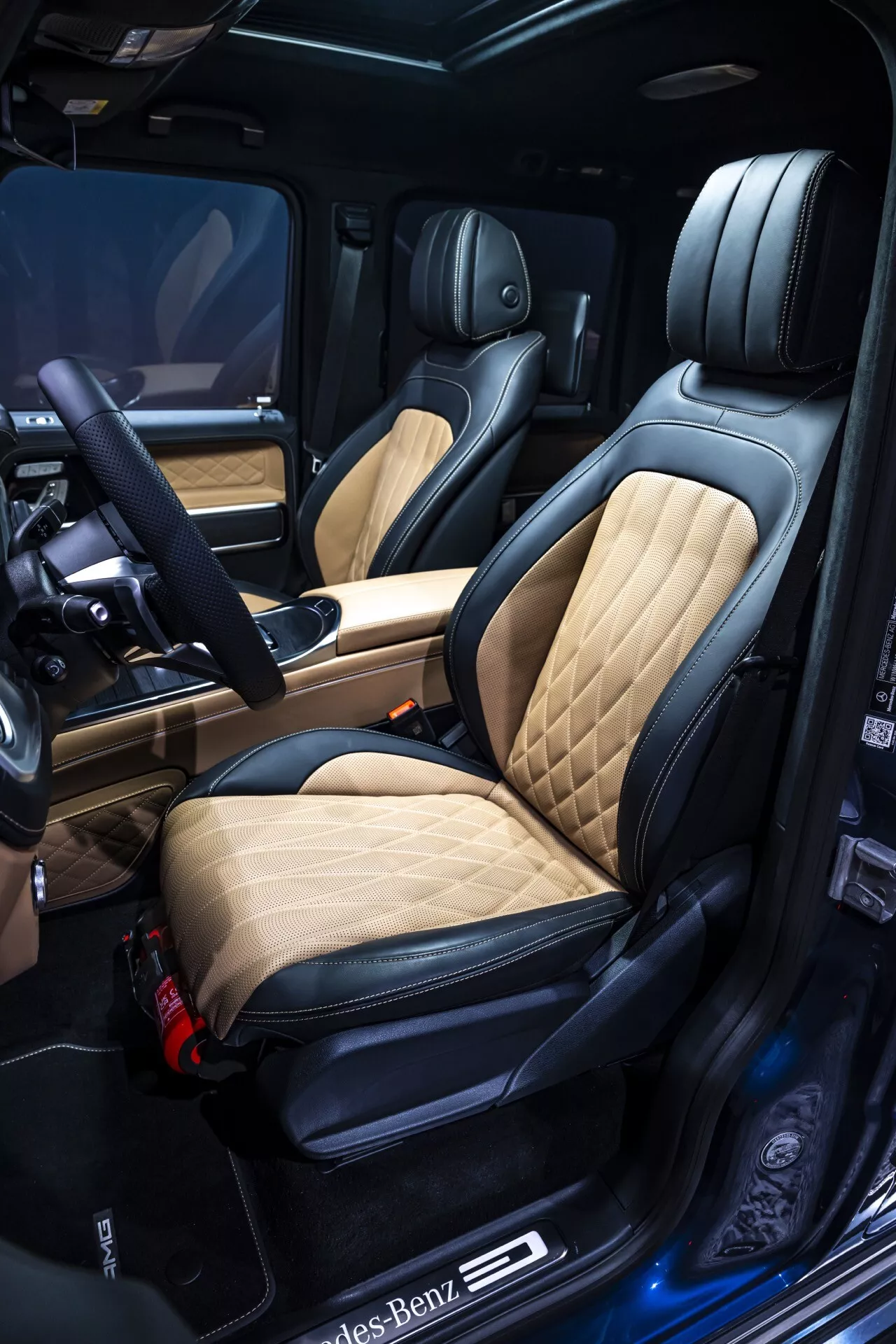 Luxusný interiér Mercedes-Benz G-Klasse 2024 s detailným pohľadom na sedadlo vodiča s prešívaným koženým poťahom v farbách tan a čierna, elektrické ovládanie sedadla s pamäťovou funkciou, osvetlený prah dverí s logom, materiály vysokej kvality a prvky pokročilej technológie