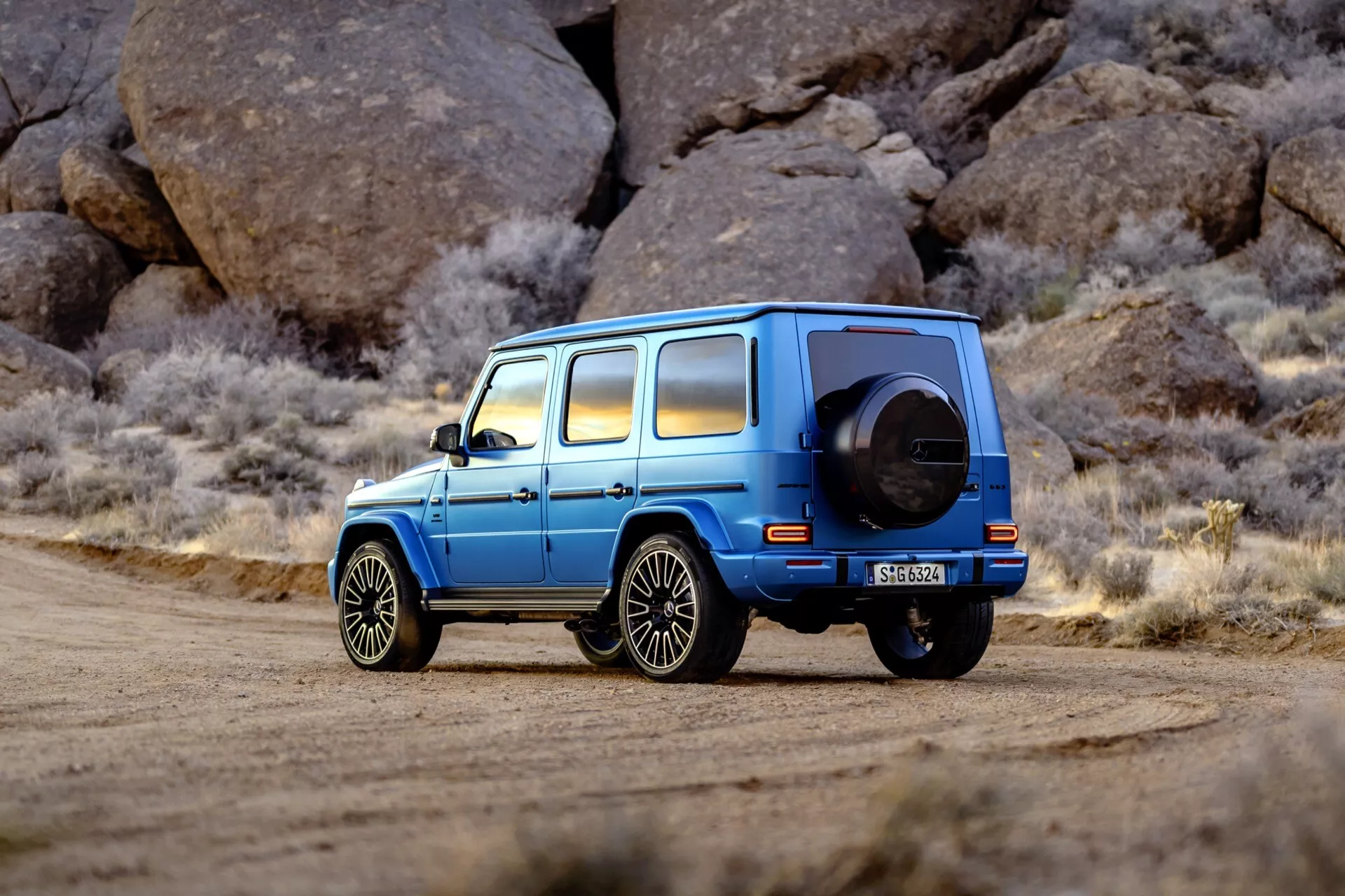 Luxusné modré SUV Mercedes-Benz triedy G (G-Klasse) 465 zaparkované na štrkovej ceste s pozadím veľkých drsných skál, zvýraznenie bočného zadného uhla, rezervného kolesa a zliatinových kolies v prostredí podobnom púšti.