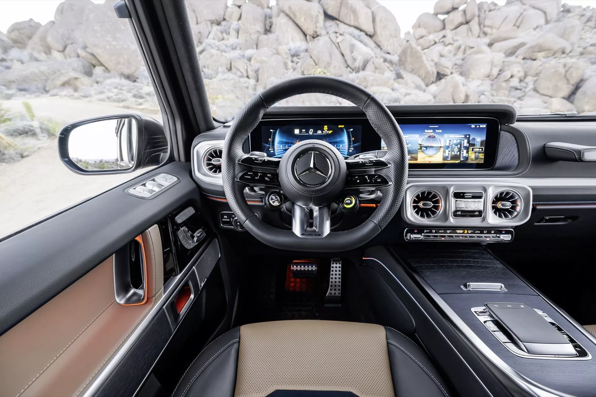 Interiér Mercedes-Benz triedy G (G-Klasse) 2024 z pohľadu vodiča s kožou potiahnutým multifunkčným volantom, digitálnym prístrojovým panelom, širokou obrazovkou informačno-zábavného systému s navigáciou a luxusným interiérom v odtieňoch tan a čierna s akcentmi z brúseného kovu a možným drevovým dekorom