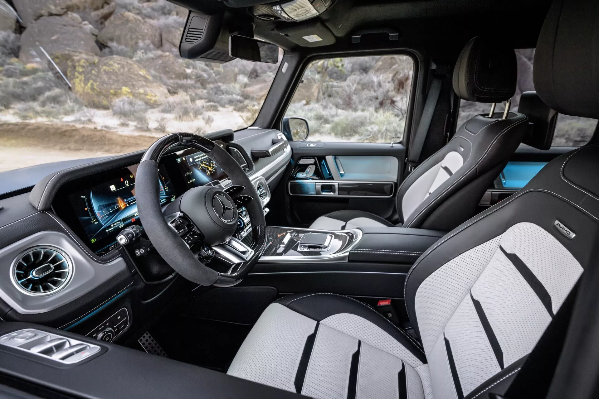 Interiér Mercedes-Benz triedy G (G-Klasse) 2024 z pohľadu spolujazdca zdôrazňujúci priestrannú kabinu s širokouhlým pohľadom, moderný multifunkčný volant, digitálny prístrojový panel, dotykovú obrazovku infotainmentu s navigáciou GPS, automatický systém riadenia klímy, vzduchové ventily v dizajne turbíny, prémiové kožené čalunenie s kontrastným šitím a športové predné sedadlá s bočným držaním