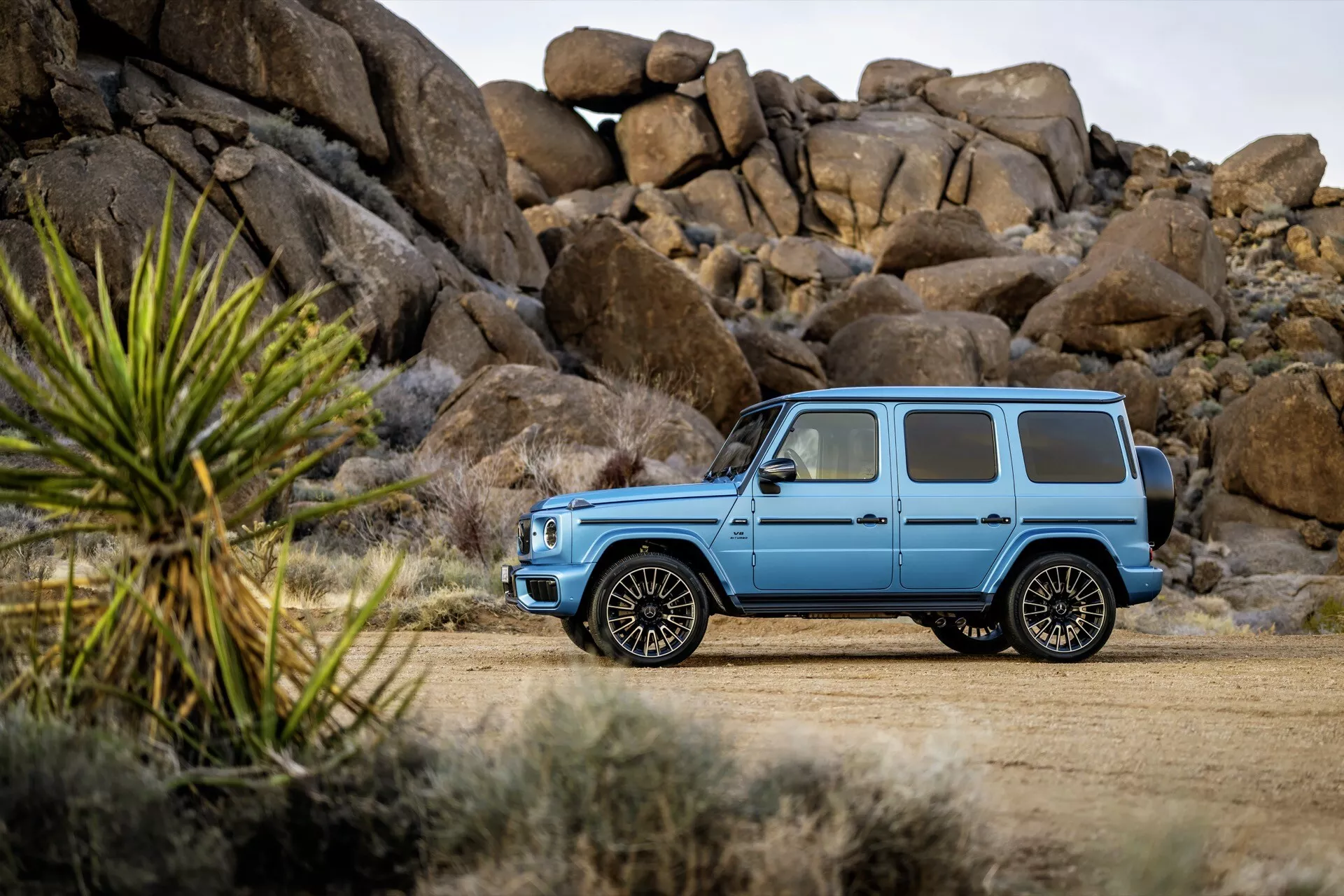 Luxusné SUV Mercedes-Benz triedy G (G 465) ročník 2024 v svetlo matnej modrej farbe, ukazuje jeho robustný dizajn a vysokú svetlú výšku na prašnej ceste, s detailne spracovanými zliatinovými kolesami a oknami s ochranou súkromia na pozadí drsnej krajiny.