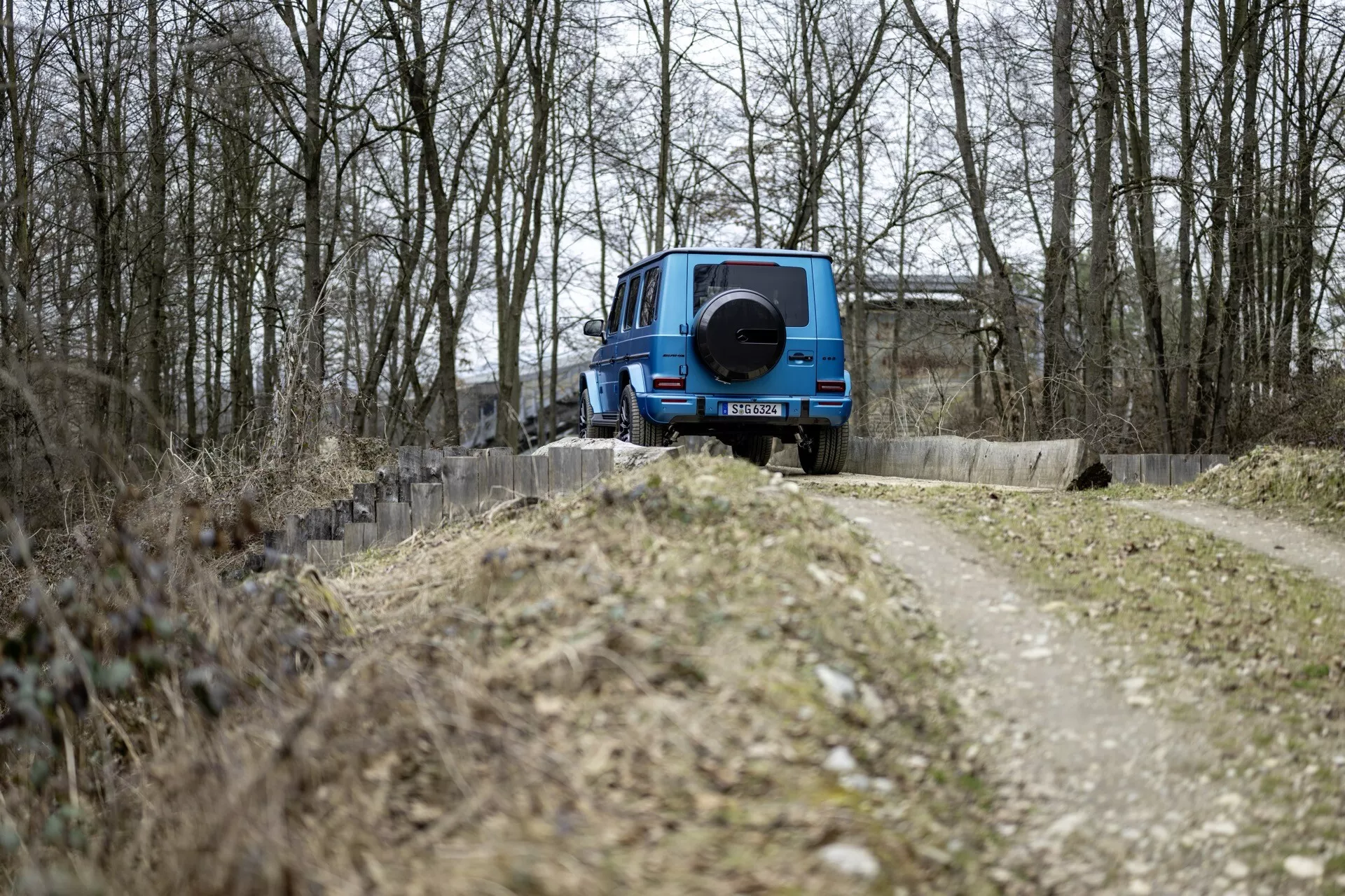 Modré SUV Mercedes-Benz triedy G 2024, otočené zadnou časťou k fotoaparátu vedľa úzkej poľnej cesty medzi opadanými stromami, ukazuje dizajn pre jazdu v teréne so záložným kolom na zadnej časti, pri zamračenom osvetlení bez viditeľných technologických prvkov na zadnom exteriéri.