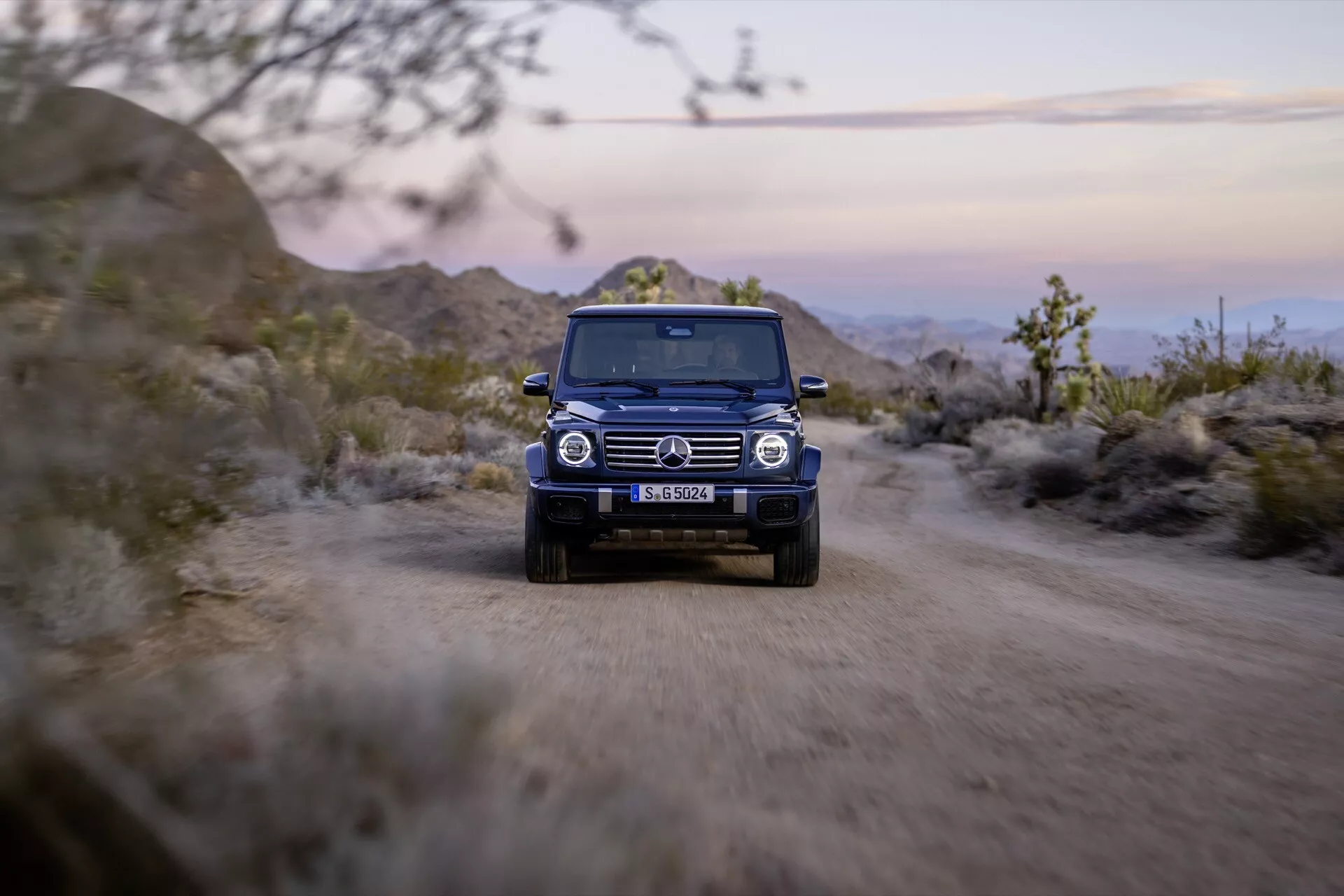 SUV Mercedes-Benz triedy G 2024 idúce po nezpevnenej ceste so zapnutými svetlometmi, čo zdôrazňuje jeho robustný dizajn a schopnosti v teréne z pohľadu spredu pri súmraku
