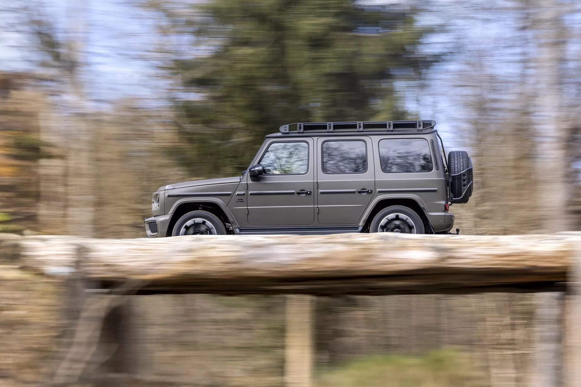 Dynamický bočný profil SUV Mercedes-Benz triedy G (G-Klasse) 2024 ukazujúci jeho pohyb s rozmazaným prírodným pozadím, zdôrazňujúci jeho štvorcový dizajn, strešný nosič a zliatinové kolesá