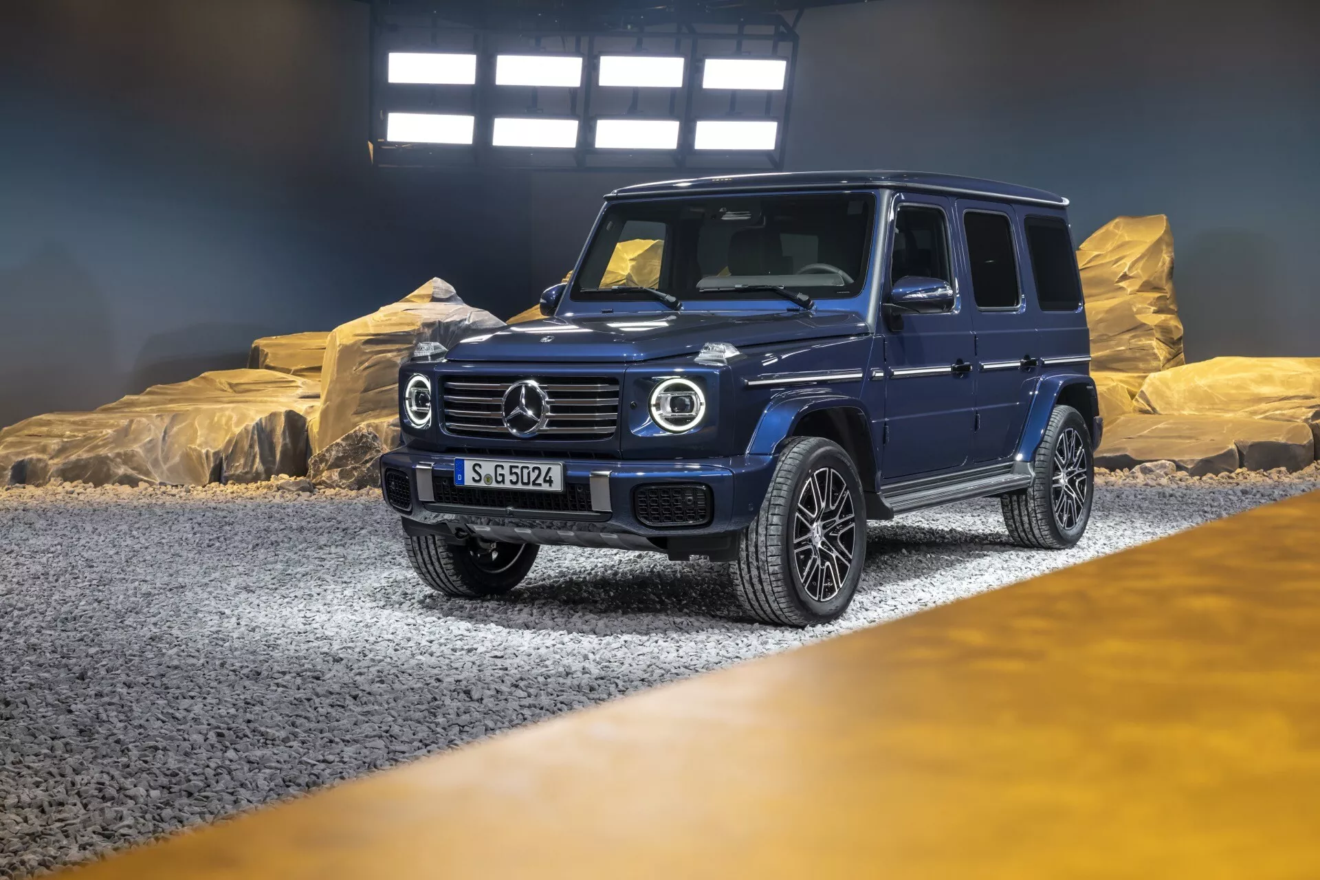 Luxusné SUV Mercedes-Benz triedy G (G-Klasse) pre rok 2024 v showroom-e s dramatickým osvetlením, zobrazujúce diagonálny pohľad zpredu a z boku, ktorý zdôrazňuje jeho kruhové LED svetlomety, výraznú mriežku s veľkým emblémom, robustný predný nárazník s pokročilou technológiou, viacramenné zliatinové kolesá a odolný, no sofistikovaný dizajn vhodný pre terénne aj mestské použitie, s evidenčnou značkou 'S-GO524'.