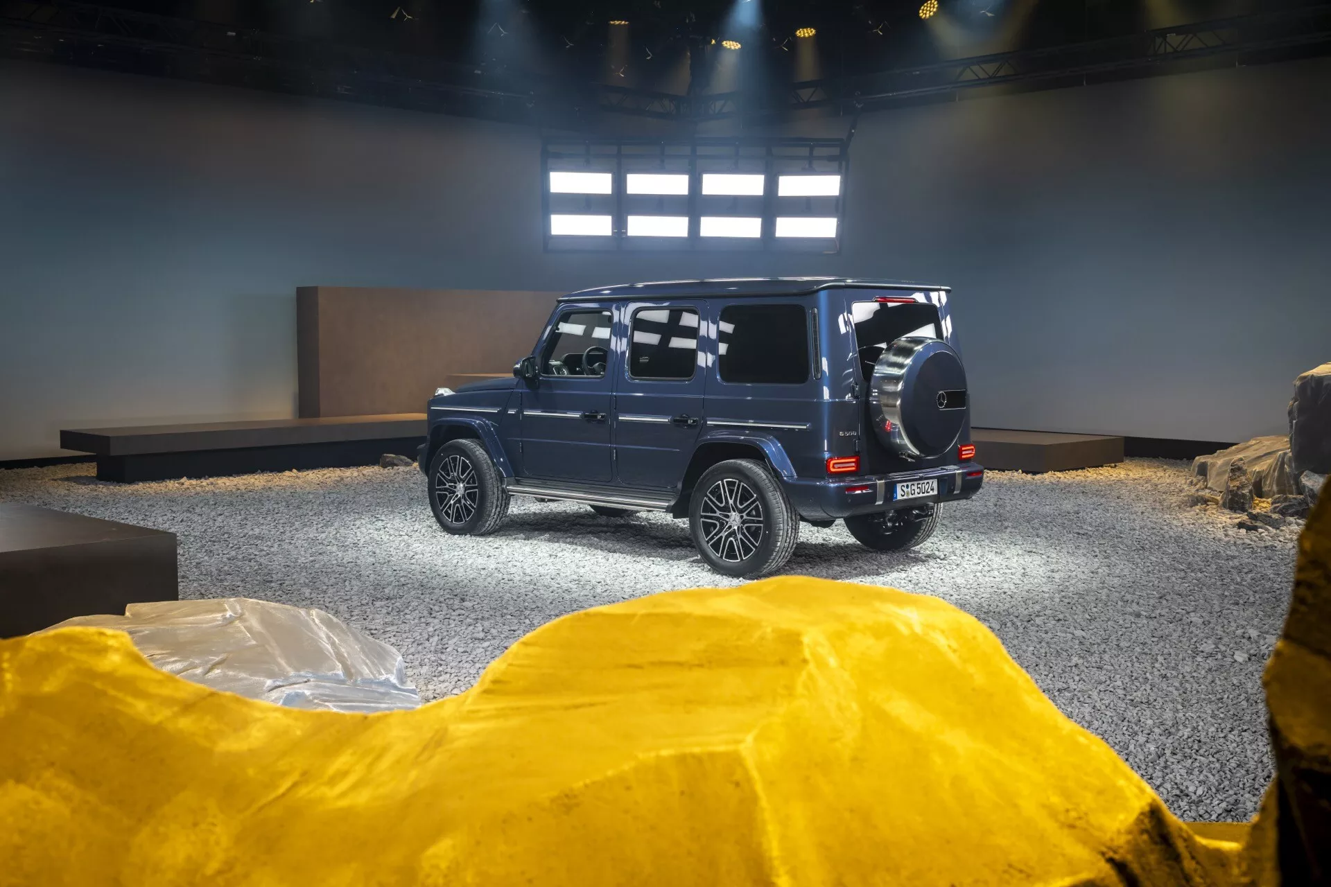 SUV Mercedes-Benz triedy G (G-Klasse) 2024 v showroome, s pohľadom na zadnú a bočnú stranu s vyzdvihnutými off-road schopnosťami vďaka náhradnému kolesu na zadnej strane, zliatinovým kolieskam a vysokej svetlej výške na aranžovanom pozadí