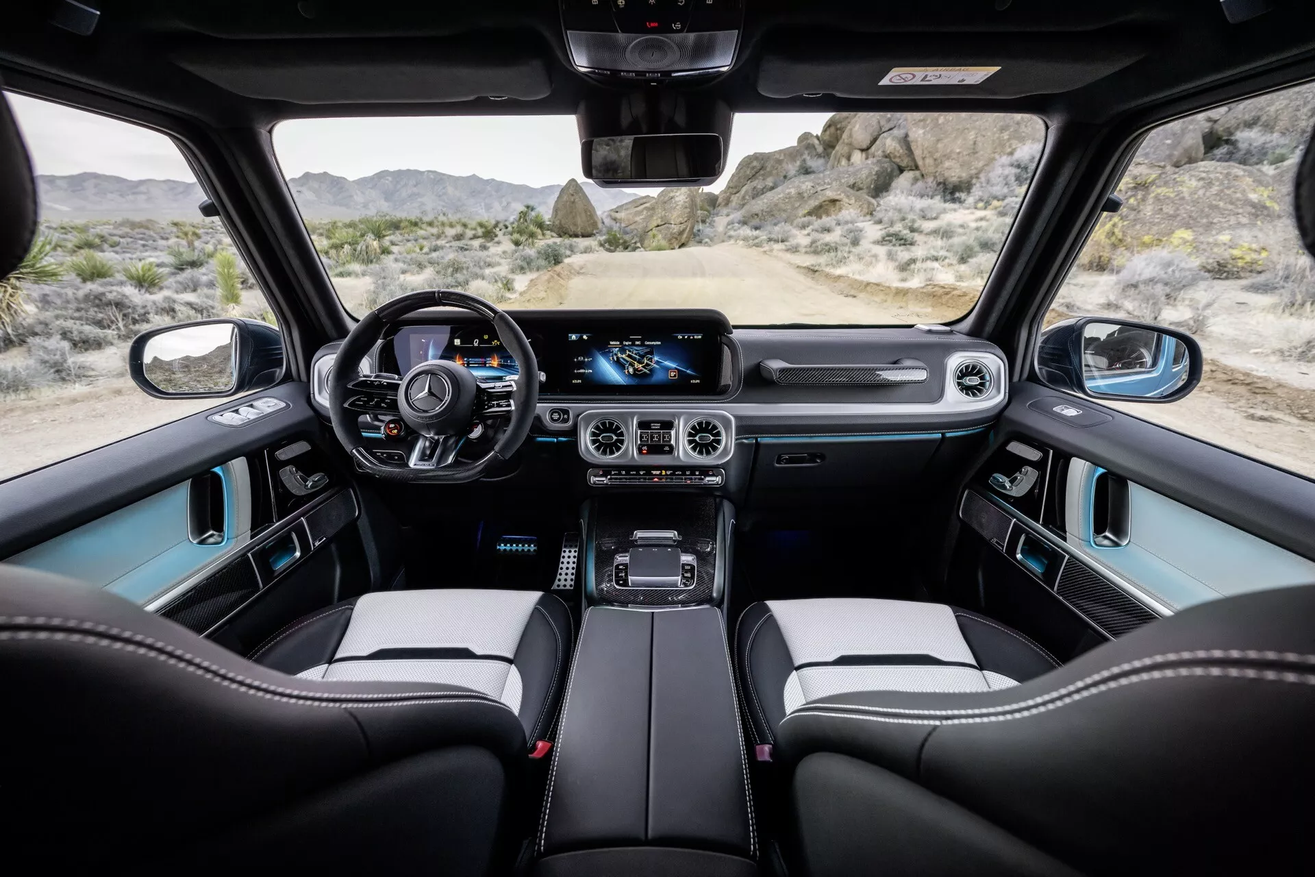 Pohľad na interiér Mercedes-Benz triedy G 2024 zo zadného sedadla, zobrazujúci palubnú dosku, digitálny prístrojový panel, volant obalený kožou s ovládacími prvkami, veľký dotykový infotainment systém, dvojzónovú klimatizáciu, robustnú radicu páku, predné sedadlá z prémiových materiálov a panoramatické strešné okno.