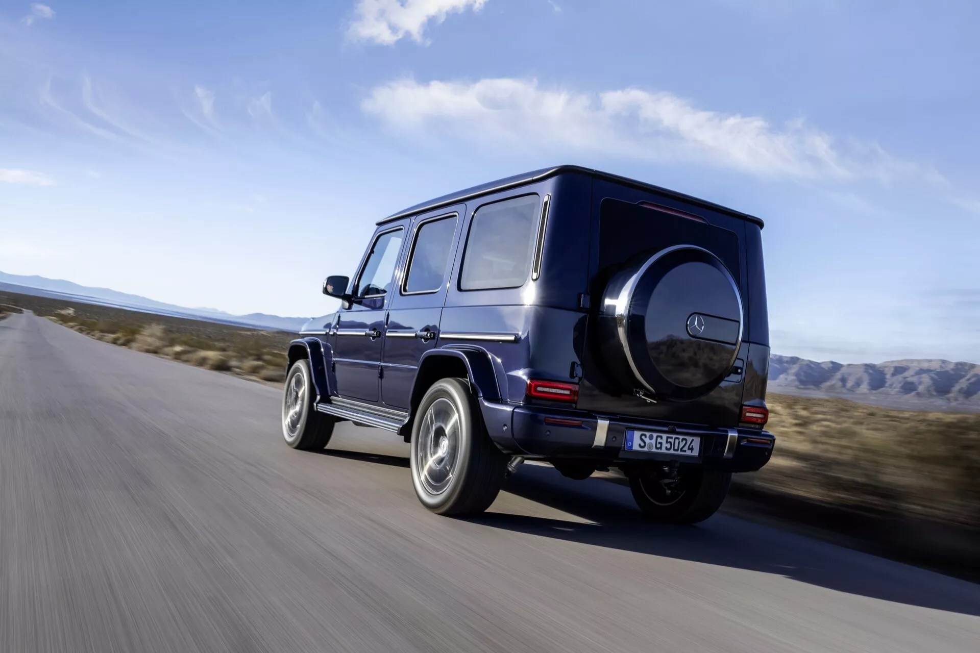 Luxusné SUV Mercedes-Benz G-Klasse 2024 tmavé farby idúce po asfaltovej ceste v prostredí pripomínajúcom púšť, predvádzajúce svoj pohľad na zadnú štvrtinu s lesklým ráfikom, kockatým dizajnom a off-roadovými prvkami ako je rezervné koleso a LED zadné svetlá.