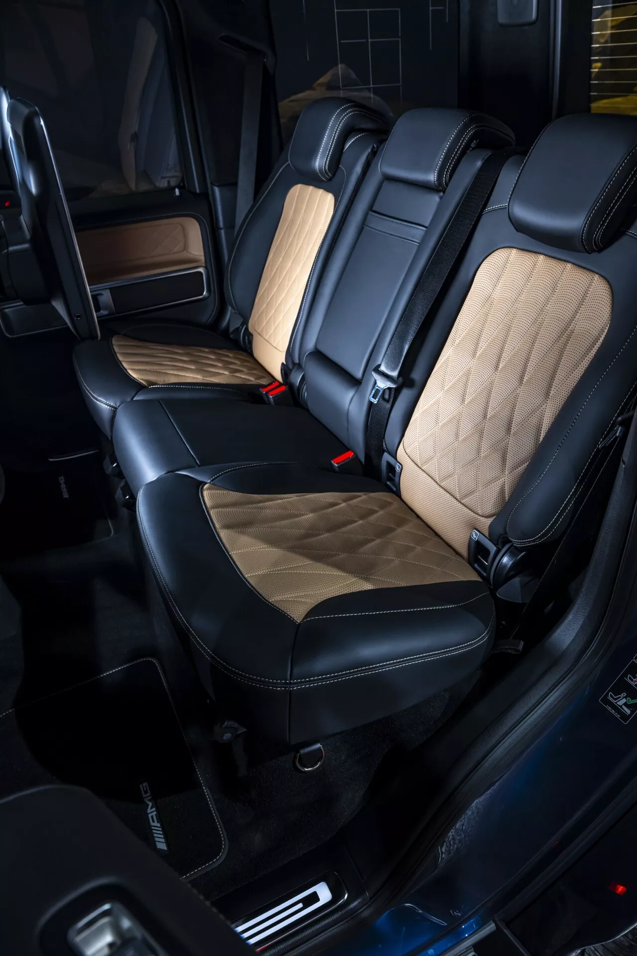 Zadný cestujúci priestor Mercedes-Benz G-Klasse (G 465) pre rok 2024 predstavujúci prémiové sedadlá v čiernej a svetlo béžovej farbe s diamantovým prešívaním, červené spony bezpečnostných pásov, dverné panely s drevotrieskovou úpravou a pokročilé funkcie kabíny, ktoré zvýrazňujú luxusný interiér a pohodlie.