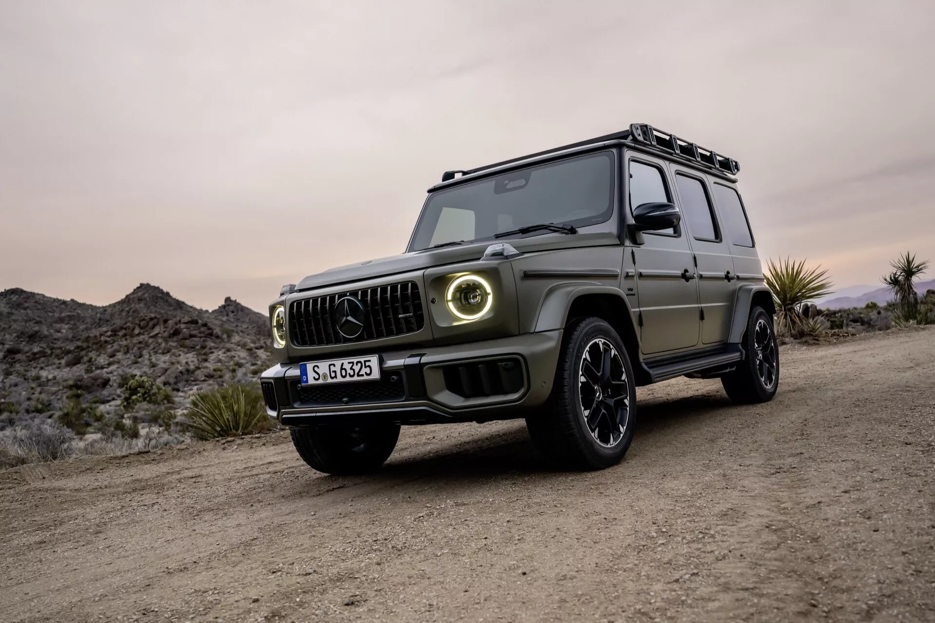 Luxusné SUV Mercedes-Benz triedy G 2024 vo výhľade z tri štvrtiny spredu, prezentujúci moderný dizajn, LED svetlomety a schopnosti v teréne na nezpevnenom povrchu