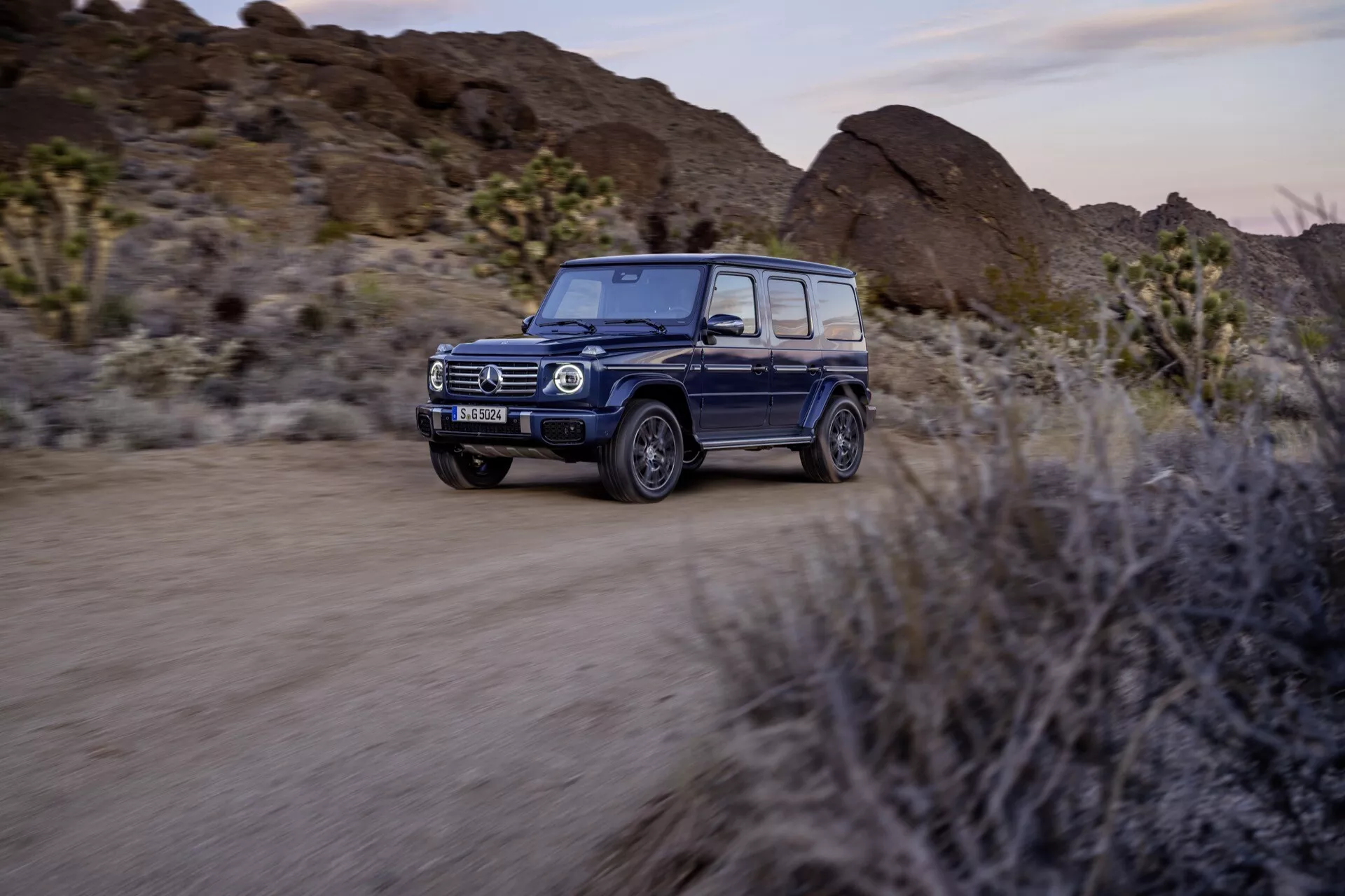 SUV Mercedes-Benz triedy G 2024 idúce po nezpevnenej púštnej ceste, s LED svetlometmi, adaptívnou technológiou, luxusným interiérom, dizajnom vhodným do terénu a pokročilými bezpečnostnými funkciami s rozmazaním v pohybe na zdôraznenie dynamických schopností