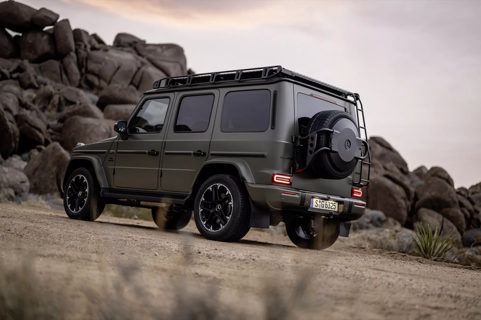 Mercedes-Benz trieda G (G-Klasse 465) 2024 v matnej olivovo zelenej farbe, prezentujúci svoj bočný a zadný pohľad s funkciami pre jazdu v teréne, umiestnený na prašnej ceste proti pozadiu skalnatej krajiny.