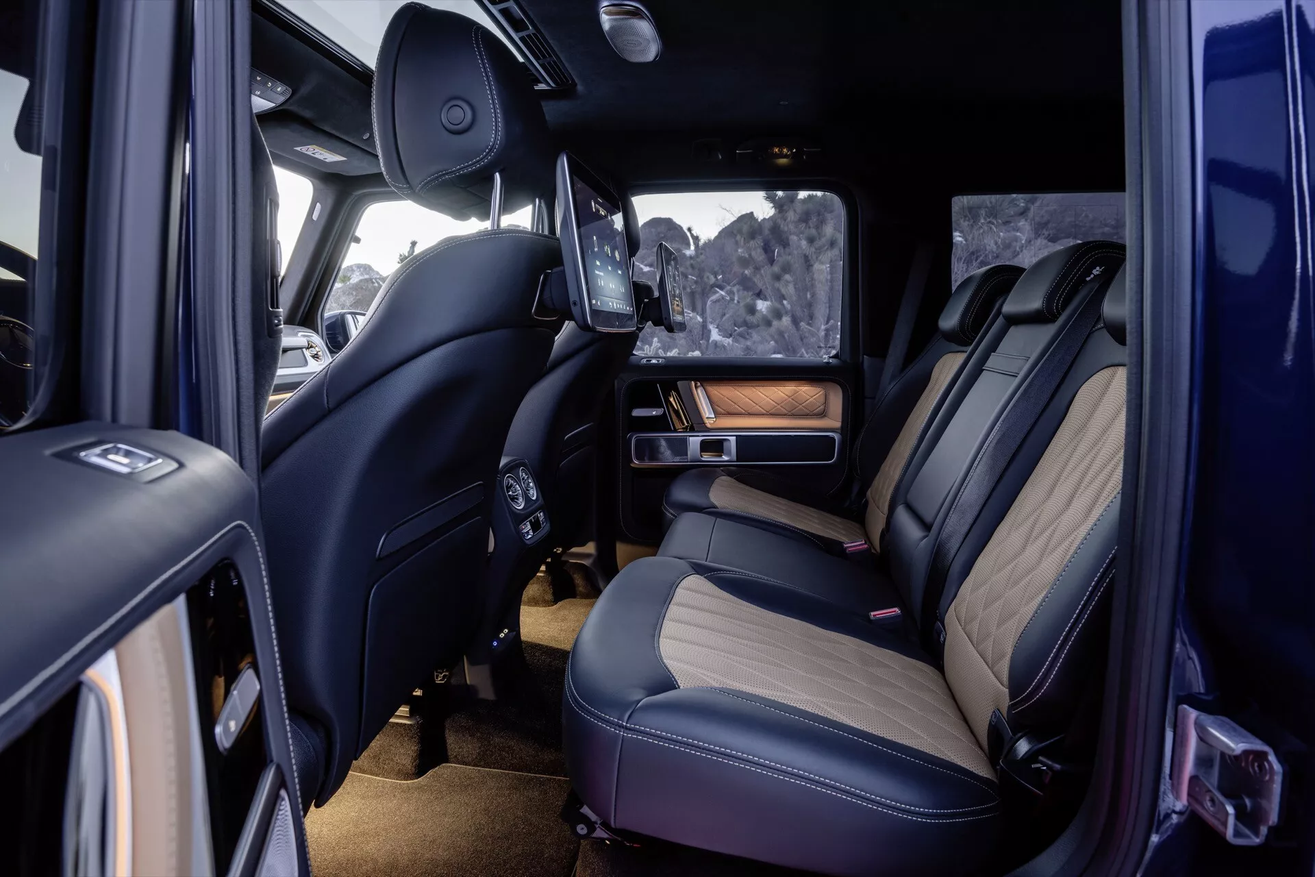 Interiérový pohľad na Mercedes-Benz triedy G 465 pre rok 2024 predstavujúci prémiové zadné sedenie, detailné šitie, luxusné materiály s drevitými doplnkami, pokročilé technologické funkcie vrátane zasúvateľnej obrazovky zábavného systému, ambientného osvetlenia a klimatizácie z perspektívy otvorených dverí spolujazdca