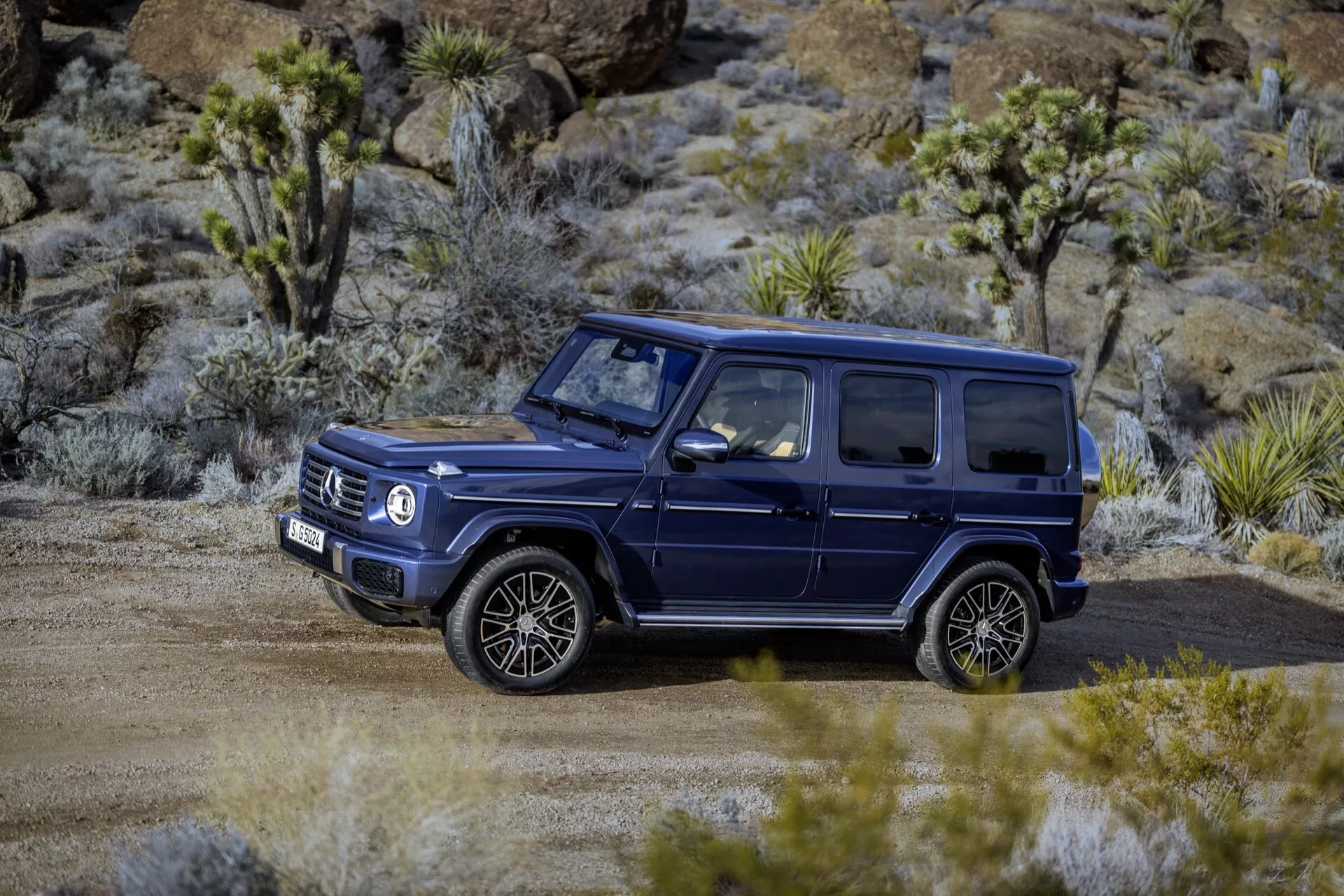 Mercedes-Benz triedy G 465 z roku 2024, tmavé SUV zaparkované v teréne na nezpevnenej ceste so suchým rastlinstvom, ukazujúce jeho robustný, štvorcový dizajn, veľké viacramenné kolesá a nosiče na streche vhodné pre dobrodružstvá a luxusné off-road schopnosti.