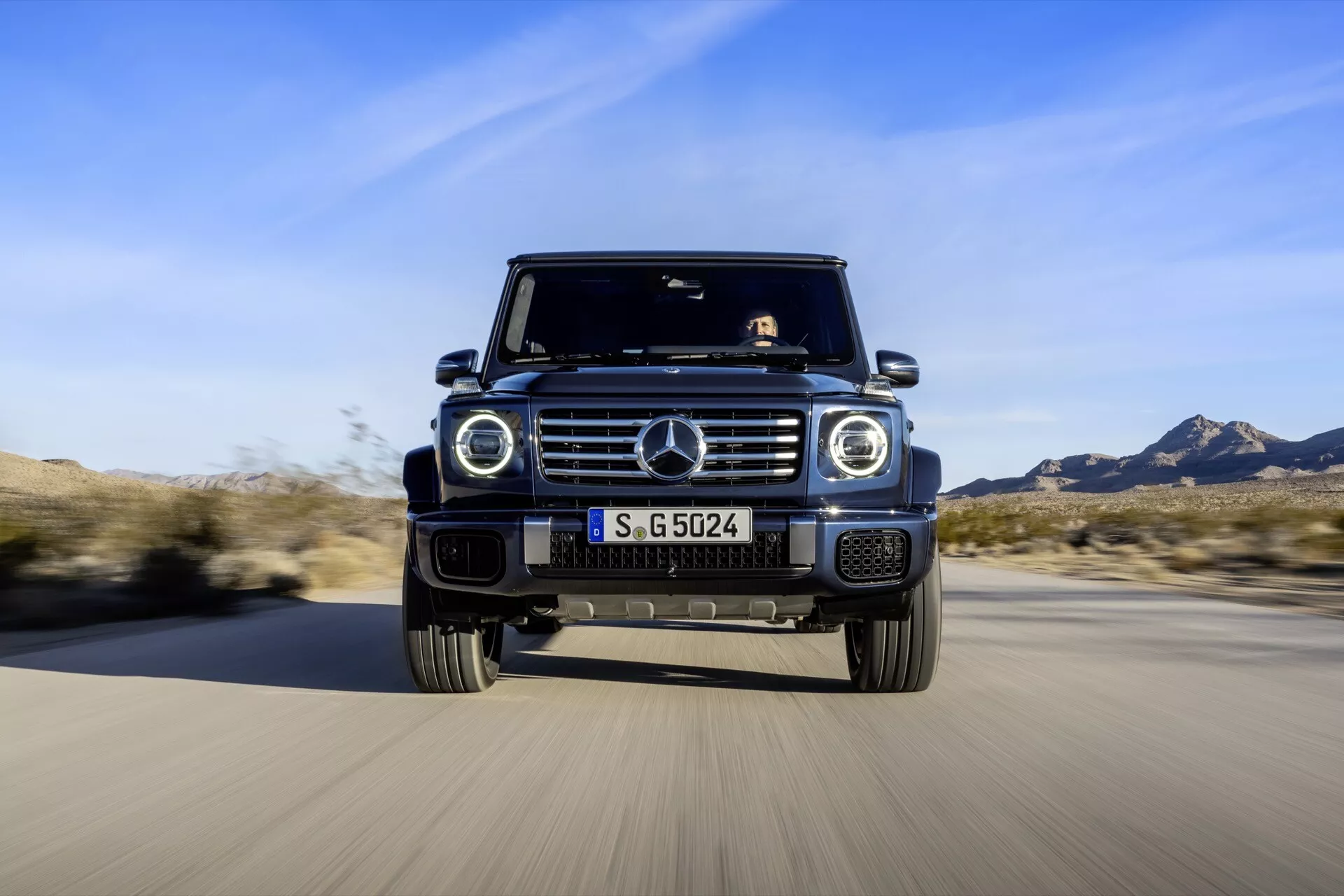 SUV Mercedes-Benz triedy G 2024 sa blíži na ceste, ukazujúc nízky uhol pohľadu na jeho emblematickú mriežku, kruhové LED svetlá, ochrannú dosku na prednom nárazníku, prednú kameru a jeho charakteristický škatuľový siluetu proti rozmazanému pozadiu, čo zdôrazňuje dynamickú prítomnosť a pokročilé funkcie vozidla.