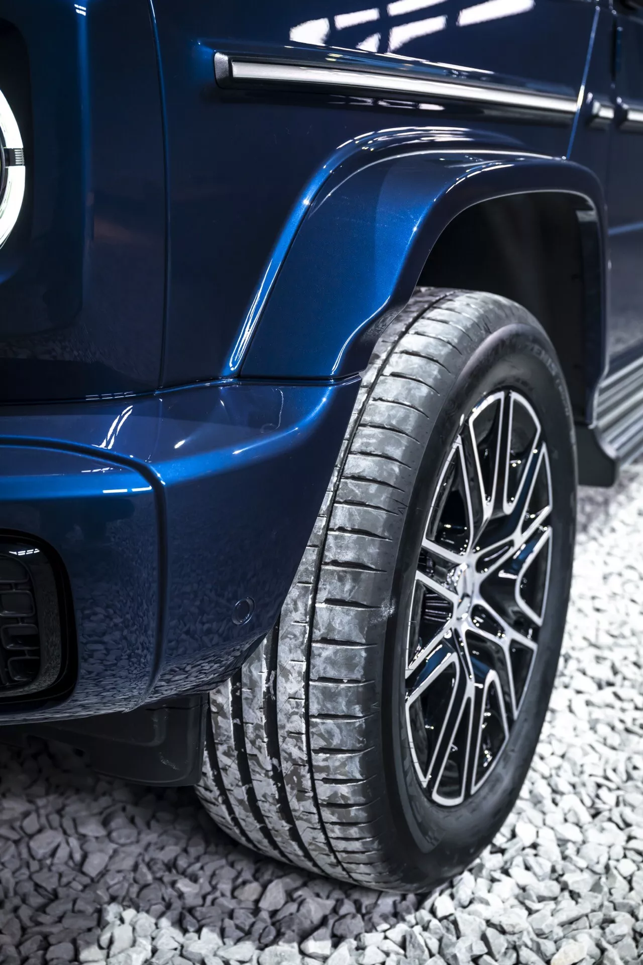 Detailný pohľad na zadné koleso a karosériu Mercedes-Benz triedy G 2024 v modrej farbe, s viacramenným zliatinovým kolom, výrazným dezénom pneumatiky a výraznými blatníkmi naznačujúcimi luxusné alebo terénne schopnosti