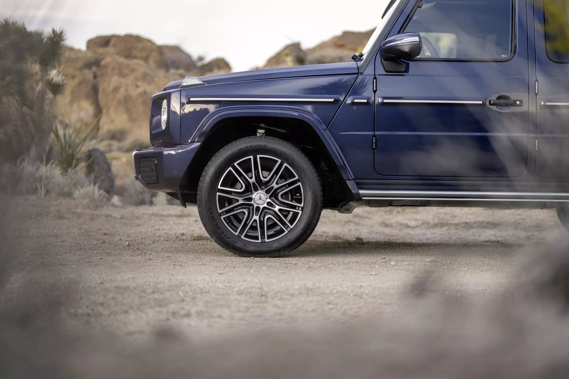 Detailný pohľad na predné ľavé koleso a blatník tmavomodrého Mercedes-Benzu G-Klasse 2024, zdôrazňujúci luxusné zliatinové kolesá a bočný profil naznačujúci terénne schopnosti