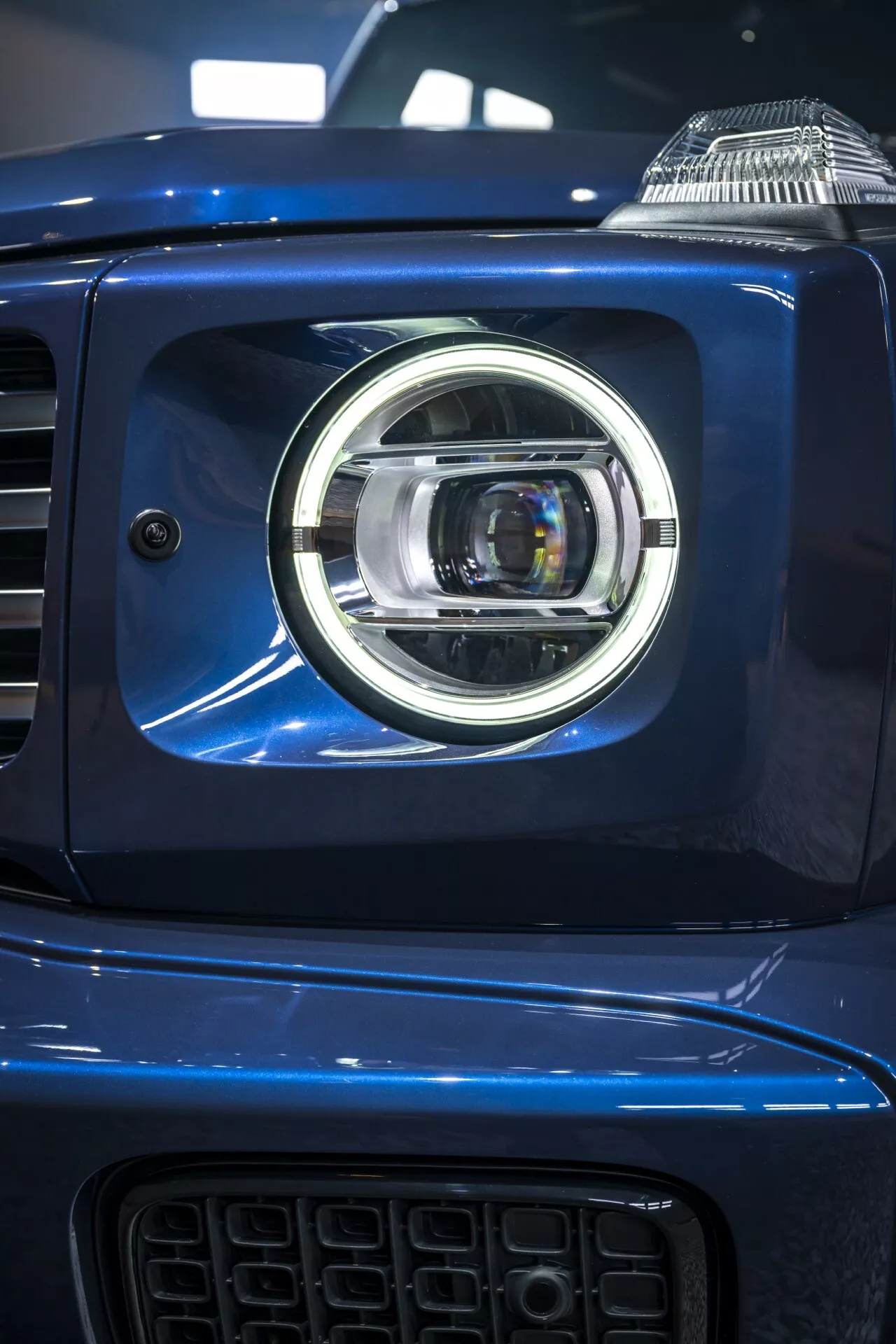 Detailný pohľad na ľavú svetlometovú súpravu Mercedes-Benz G-Klasse 2024, predvádzajúci moderný dizajn LED projektoru s chrómovými akcentmi na metalickom modrom karosérii