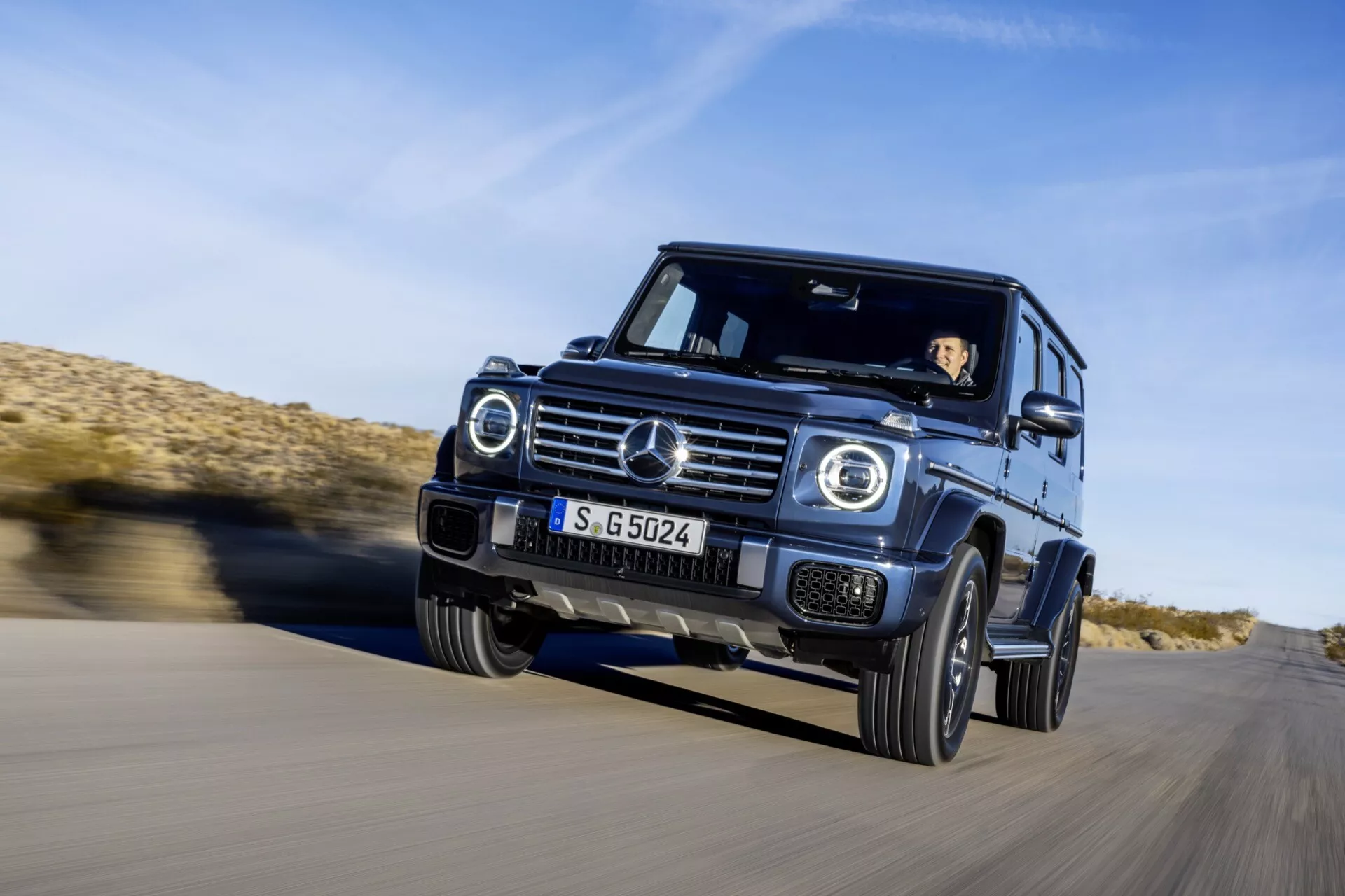 SUV Mercedes-Benz triedy G 2024 jazdiaci po asfalte v púštnej oblasti, pohľad z predného trištvrte pod úhlom zdôrazňujúci dynamický pohyb, LED svetlomety, chrómovú mriežku a schopnosti jazdy v teréne