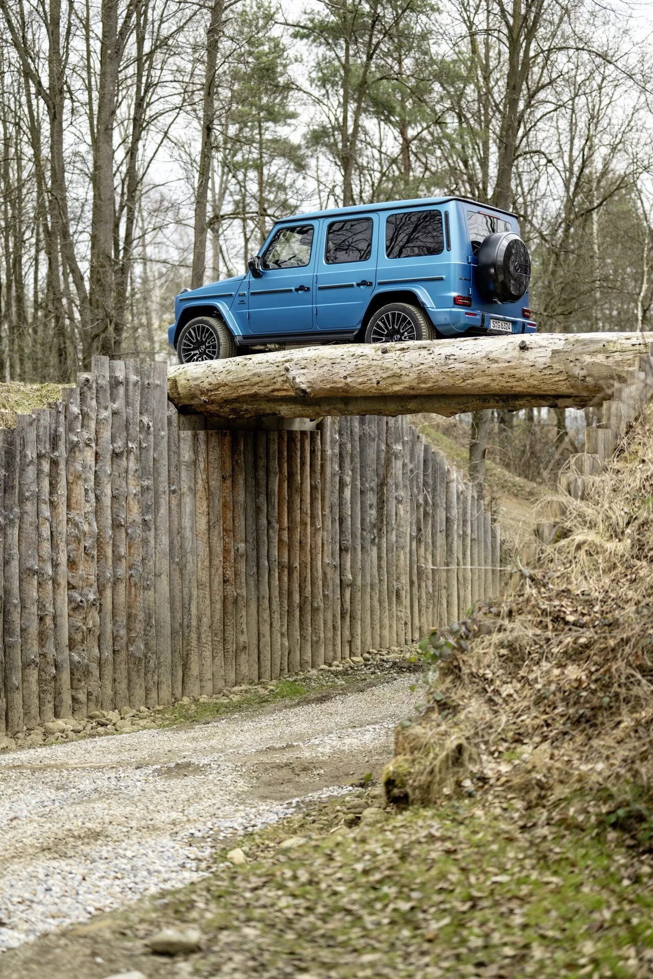 Modré SUV Mercedes-Benz G-Klasse 2024 na zdvihnutej drevenej plošine v terénnom prostredí, zobrazuje zadný a bočný pohľad s viditeľnou rezervou