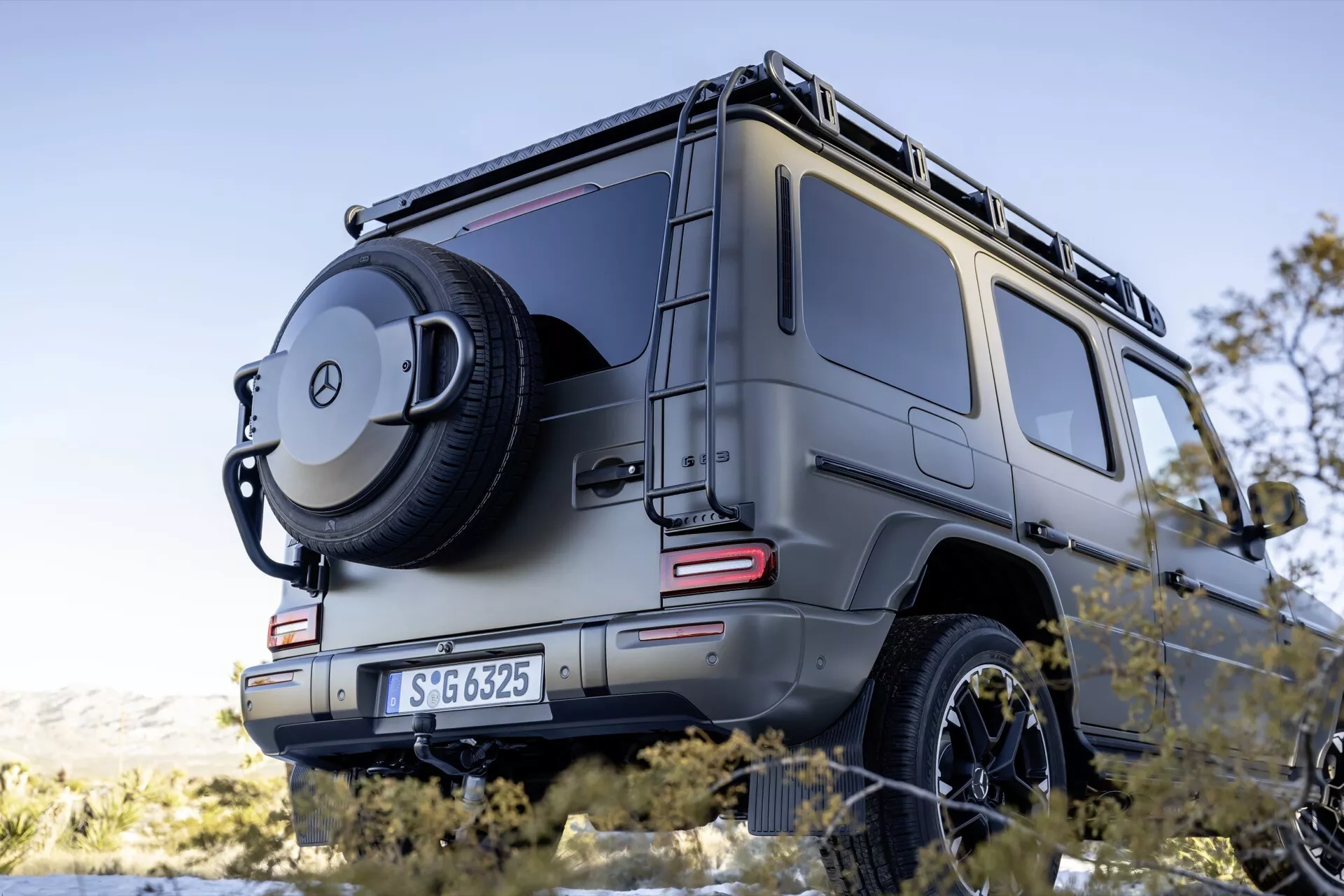 Zadný pohľad z troch štvrtín na SUV Mercedes-Benz G-Klasse 2024 ukazujúci pravú stranu vozidla z nízkeho uhla, čím zdôrazňuje jeho mohutný vzhľad s detailmi zameranými na LED zadné svetlá, rezervné koleso umiestnené na zadných dverách s krytom s emblémom, robustný štvorcový dizajn indikujúci terénne schopnosti, parkovacie senzory, ťažné zariadenie a strešný nosič na metalickej úprave pod ambientným osvetlením.