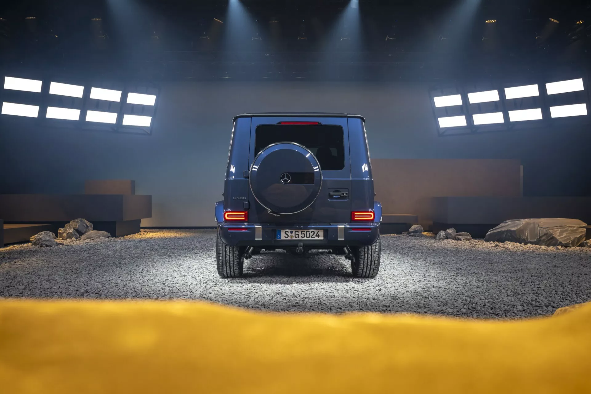 Zadný pohľad na SUV Mercedes-Benz triedy G (G-Klasse) 2024 vo falošnom terénnom prostredí v štúdiu, zdôrazňujúci výšku, zadné svetlá, rezervné koleso a strategické osvetlenie
