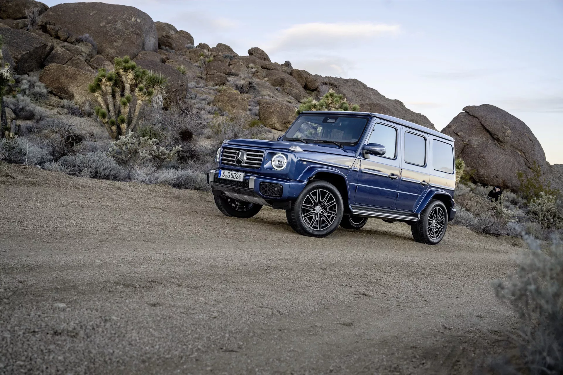 Modré SUV Mercedes-Benz triedy G 465 z roku 2024 zaparkované na drsnom teréne, ukazujúce štvorcový dizajn, vysokú svetlú výšku a prezentujúce schopnosti vozidla pre jazdu v teréne z pohľadu z troch štvrtín spredu, ktorý zdôrazňuje zliatinové kolesá a terénne pneumatiky