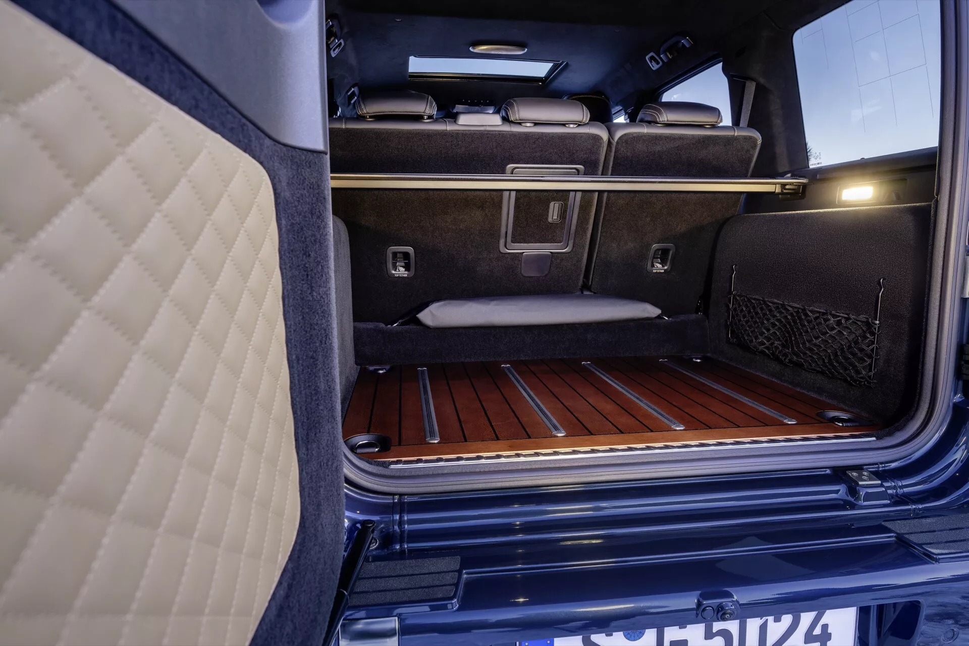 Detailný pohľad na interiér batožinového priestoru Mercedes-Benz G-Klasse 2024, zdôrazňujúci jeho prémiové kobercové čalúnenie, drevené podlahové laty, kovové kotviace úchyty pre náklad a sieťované vrecko na drobnosti, vedľa luxusných zadných sedadiel a integrovaného osvetlenia batožinového priestoru