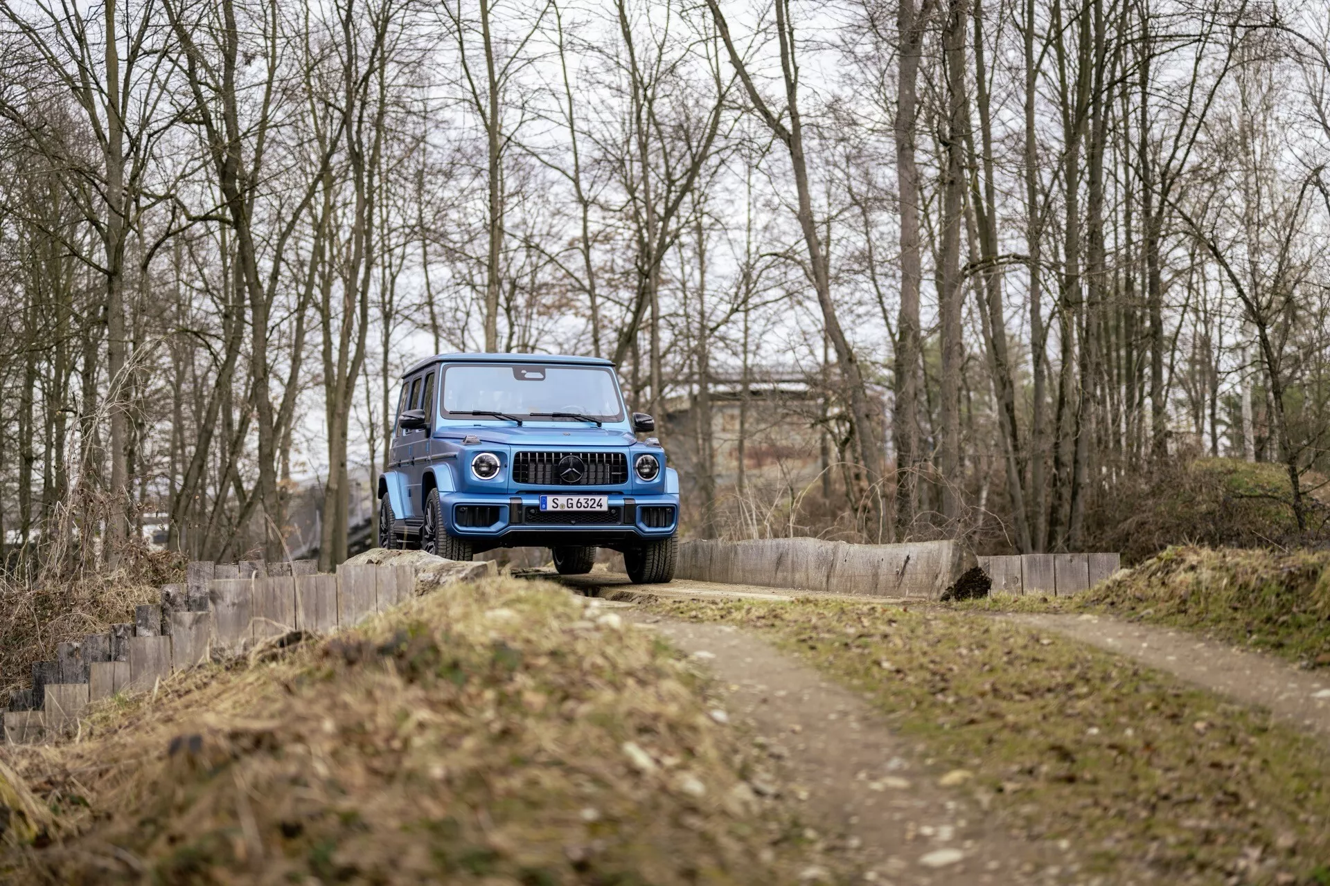 Modré terénne vozidlo Mercedes-Benz G-Klasse 2024 v pohybe na nezpevnenej ceste, pohľad z predného trojštvrtečného uhla ukazujúci prednú mriežku, kapotu, čelné sklo a vysokú svetlú výšku, v lesnom prostredí bez snehu