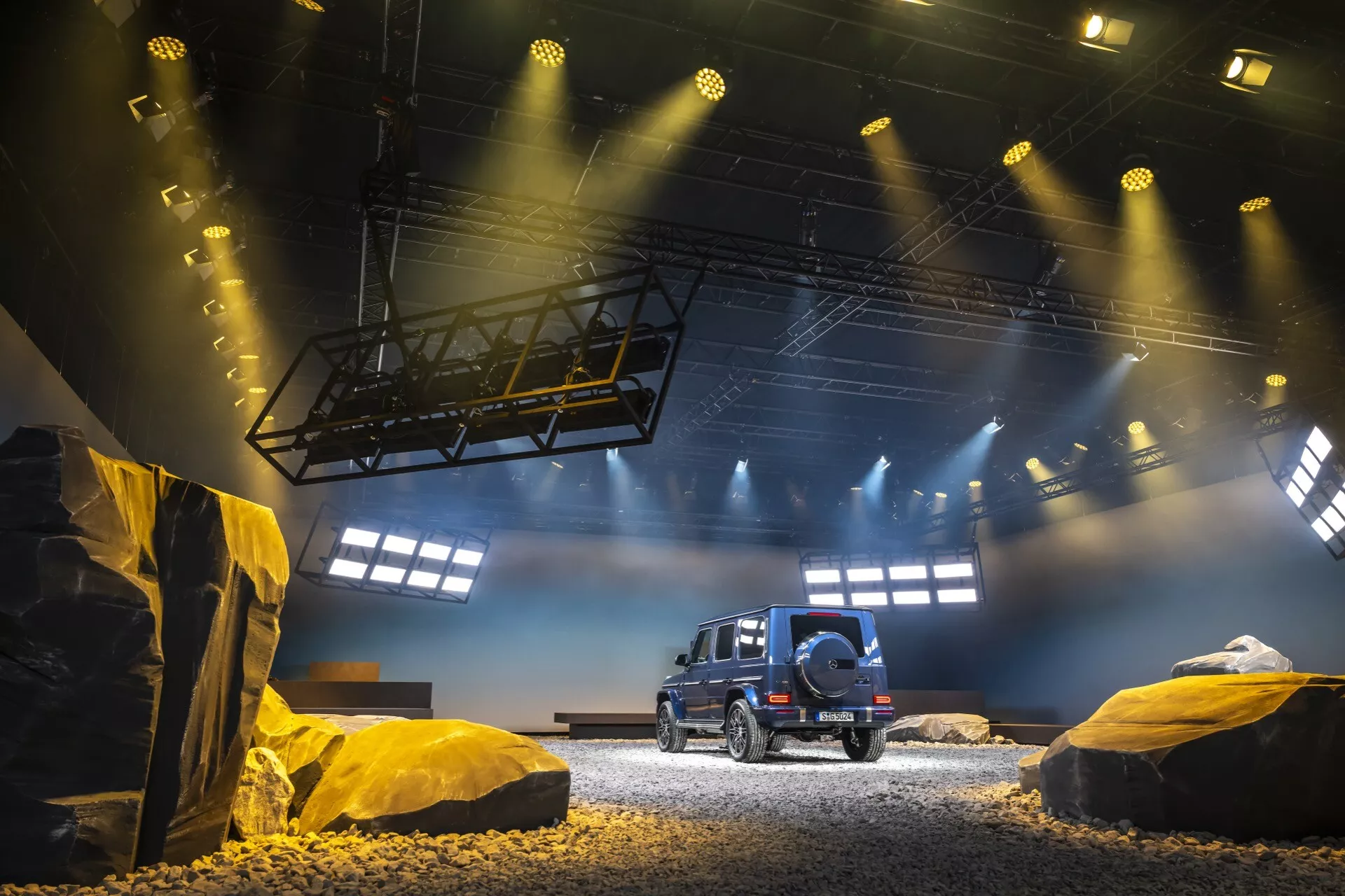 SUV Mercedes-Benz G-Klasse 2024 vystavené v simulovanom terénnom prostredí, predvádzajúce pohľad na vozidlo spredu a z boku, s detailným bočným profilom, zadným oknom, LED zadnými svetlami a rezervným kolesom umiestneným na zadných dverách pod dramatickým osvetlením výstavy