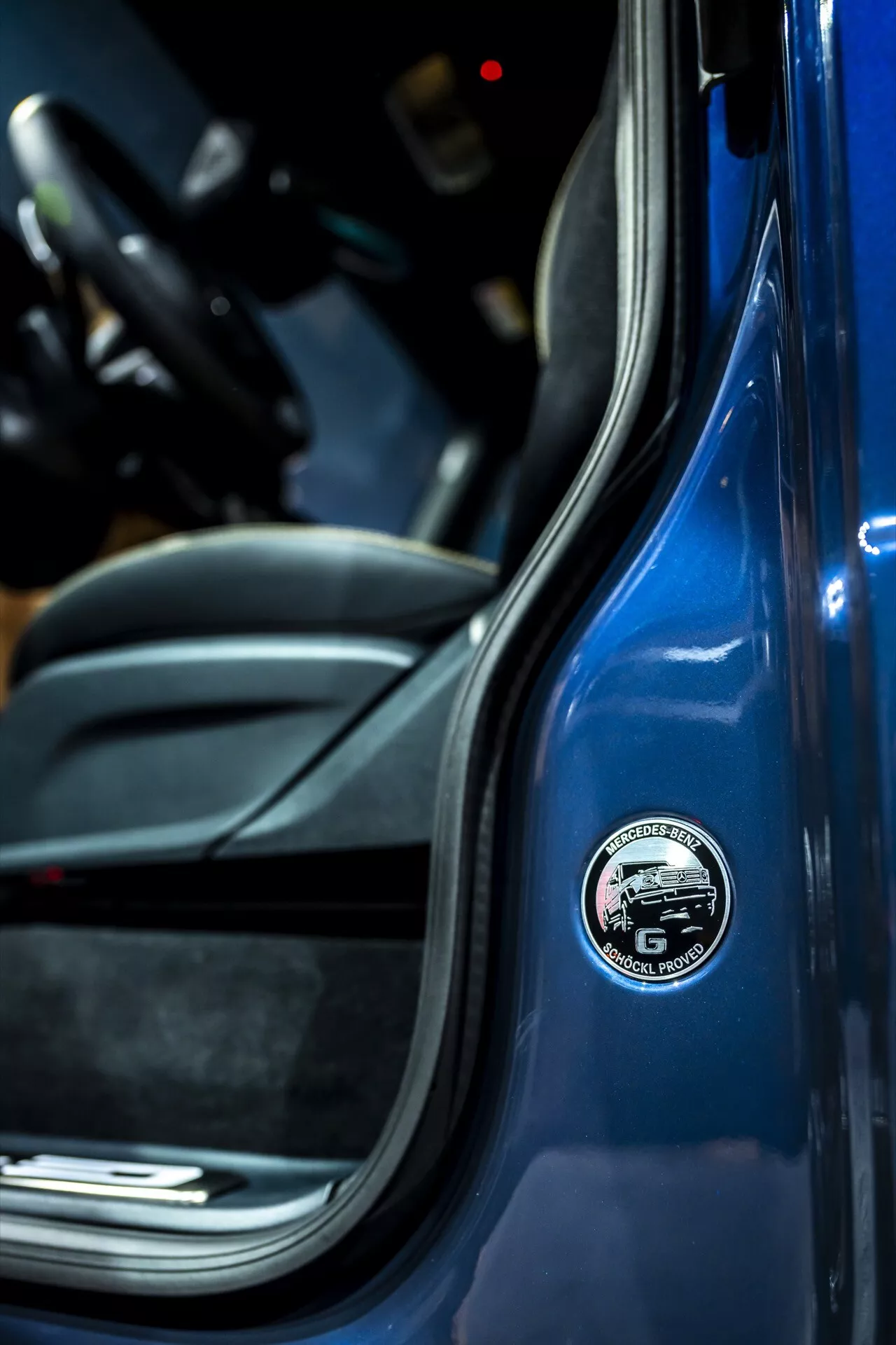 Detailný obrázok odznaku na B-stĺpiku Mercedes-Benz G-Klasse 2024 s živým modrým exteriérom a športovým čiernym interiérom, zdôrazňujúci prémiové vlastnosti vozidla so zameraním na značku a materiály vysokej kvality.