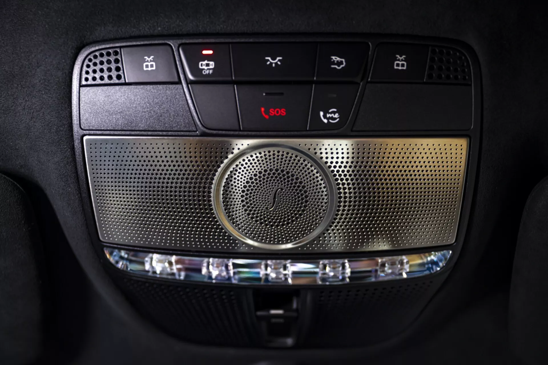 Detailný pohľad na stropnú konzolu Mercedes-Benz G-Klasse 2024 (465) s ovládacími tlačidlami, centrálnym reproduktorom, možnosťami interiérového osvetlenia, núdzovými službami a prispôsobiteľnými funkciami, zdôrazňujúci pokročilý multimediálny systém a sofistikované detaily dizajnu