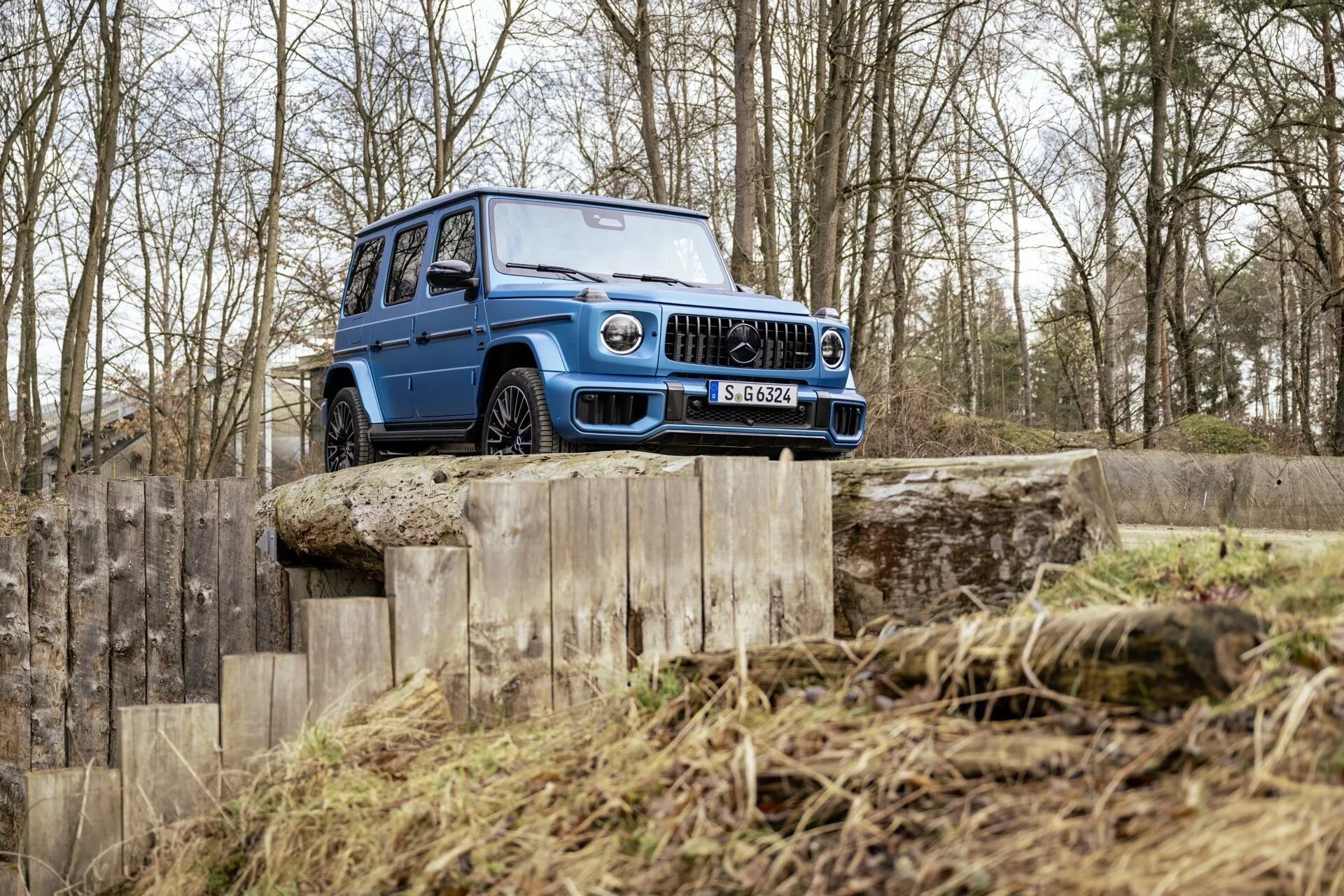SUV Mercedes-Benz G-Klasse (G-Class) 465 pre rok 2024 predvádzajúce svoje schopnosti v teréne na viacúrovňovej drevenej rampe v náročnom teréne, zdôrazňuje vysokú svetlú výšku, robustnú prednú mriežku, LED svetlomety a pneumatiky s hlbokým dezénom vhodné pre náročné použitie