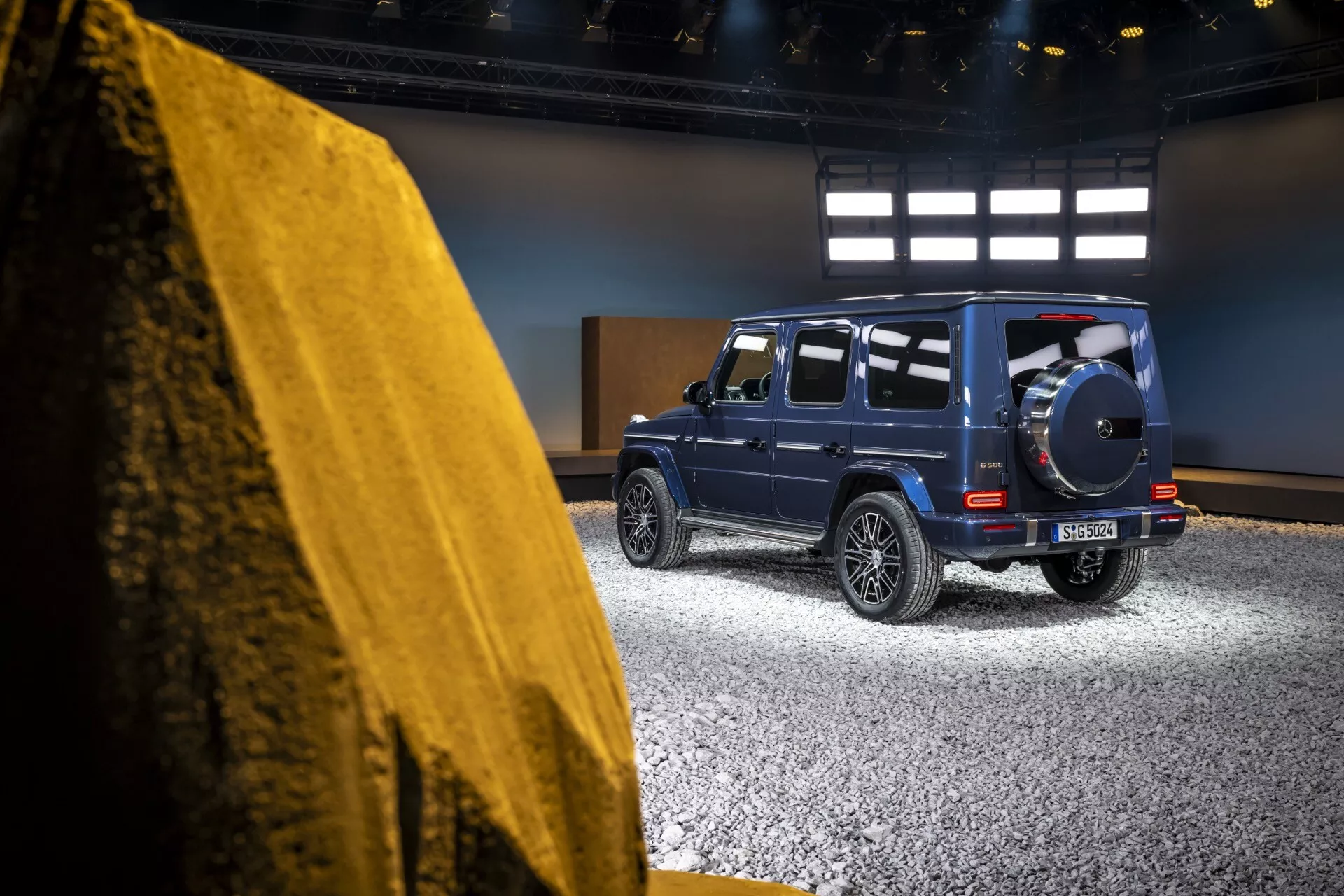 Bočný zadný pohľad na Mercedes-Benz triedy G (G 465) ročník 2024 v štúdiovom prostredí, predstavujúci jeho robustný luxusný dizajn, LED zadné svetlá a rezervné koleso na hlbokom metalickom modrom