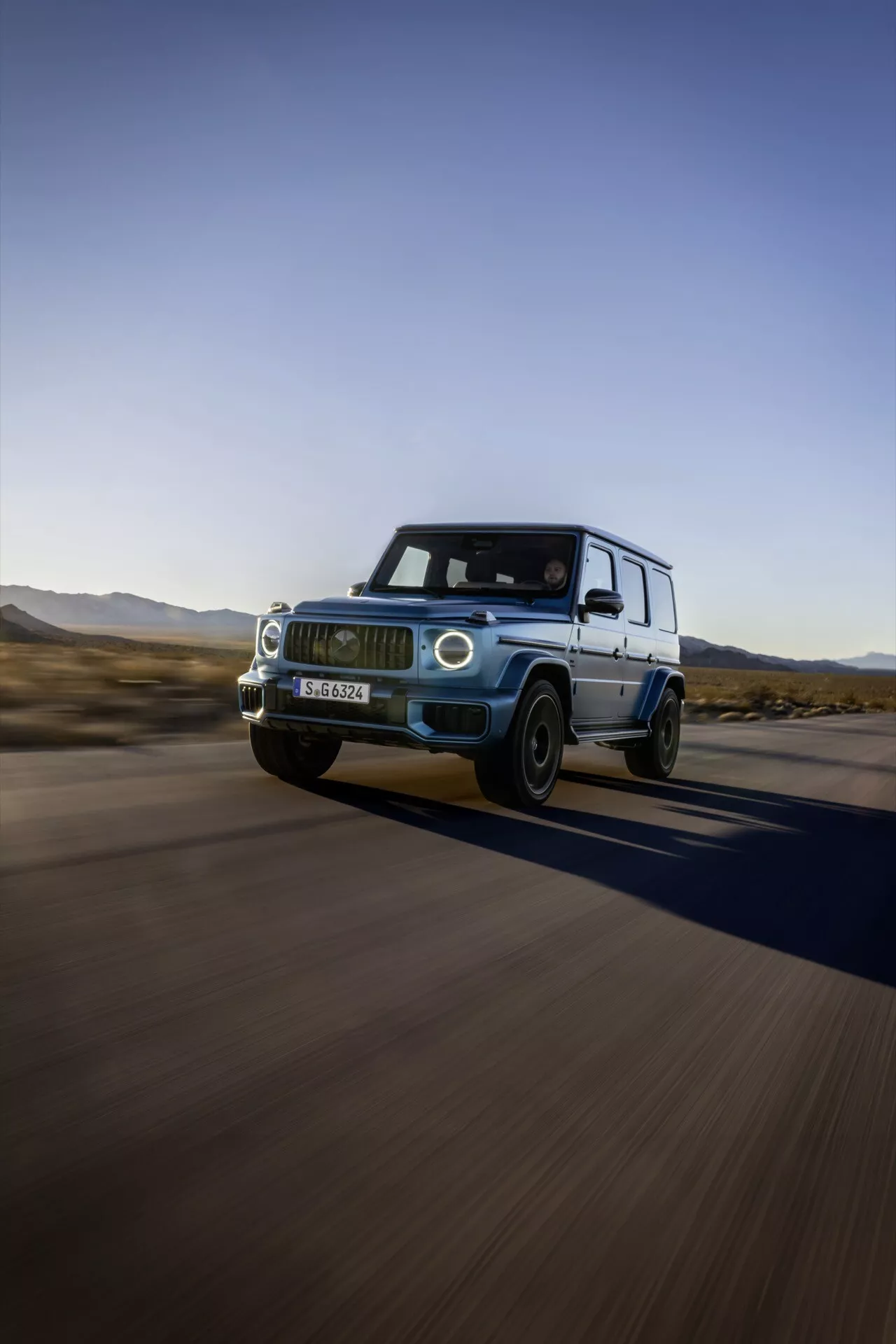Mercedes-Benz G-Klasse 2024 v pohybe, ukazuje jeho luxusný dizajn a vlastnosti z nízkeho uhla prednej a bočnej časti, zdôrazňuje LED svetlomety, prednú mriežku, zliatinové kolesá, a naznačuje jeho schopnosti vysokých rýchlostí a off-road potenciál v púštnej krajine.