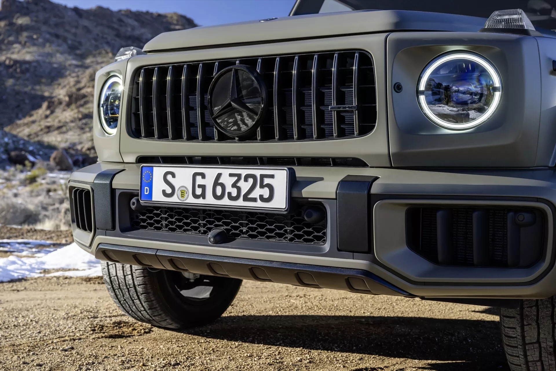 Detailný pohľad na prednú časť SUV Mercedes-Benz G-Class model 2024, zdôrazňujúci LED svetlomety, mriežku chladiča a dizajn predného nárazníka na pozadí prírody
