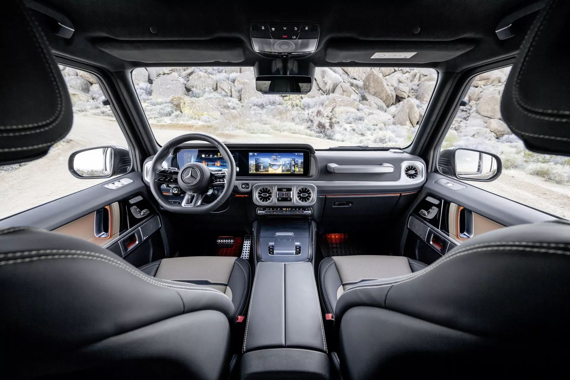 Interiér Mercedes-Benz triedy G (G-Klasse 465) z roku 2024 z pohľadu zadného sedadla ukazujúci čierne kožené sedadlá, moderný volant s ovládaním, palubnú dosku z kože a kovu, digitálny prístrojový panel, stredovú konzolu s kovovými akcentmi, veľký dotykový obrazovka infotainment systému, ovládače klimatizácie s kovovým povrchom, ovládanie osvetlenia nad hlavami, prémiový reproduktorový systém, panoramatické strešné okno, centrálnych spätné zrkadlo, a priestranné okná zvyšujúce viditeľnosť a luxusný pocit.