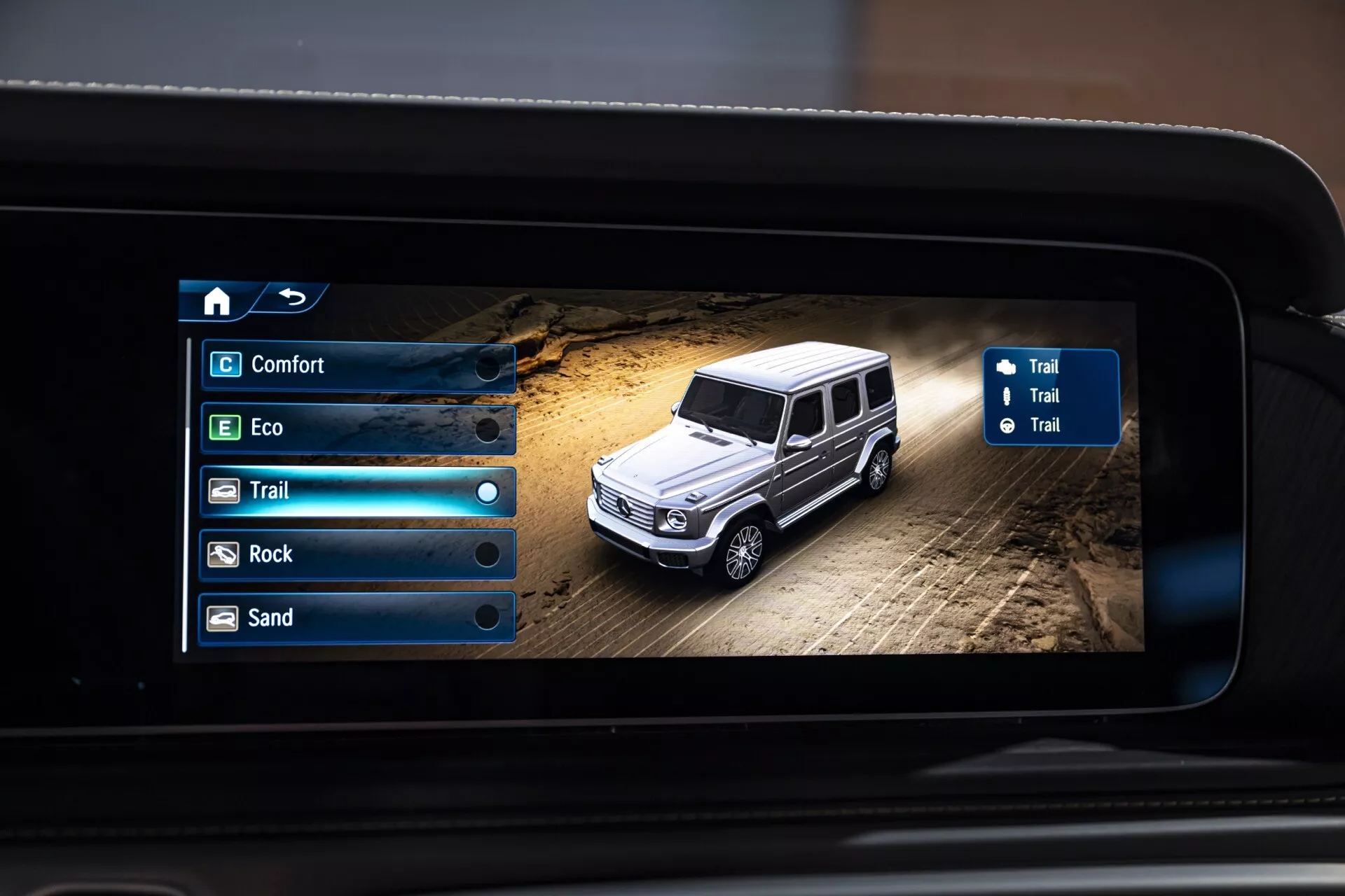 Digitálne zobrazenie Mercedes-Benz G-Klasse (trieda G) 2024 v terénnom nastavení so zvoliteľnými jazdnými režimami na obrazovke infotainment systému, pohľad na luxusné SUV z vtáčej perspektívy na prašnom teréne s navigáciou a možnosťami režimu