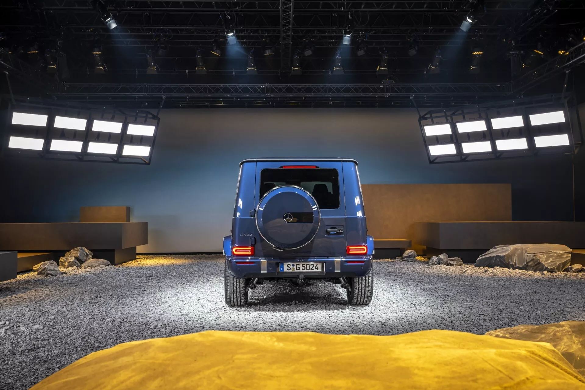 Zadný pohľad na Mercedes-Benz triedy G (G 465) ročník 2024 v inscenovanom off-road prostredí s kameňmi a texturovaným terénom, zobrazuje LED zadné svetlá, náhradné koleso a lesklý povrch.