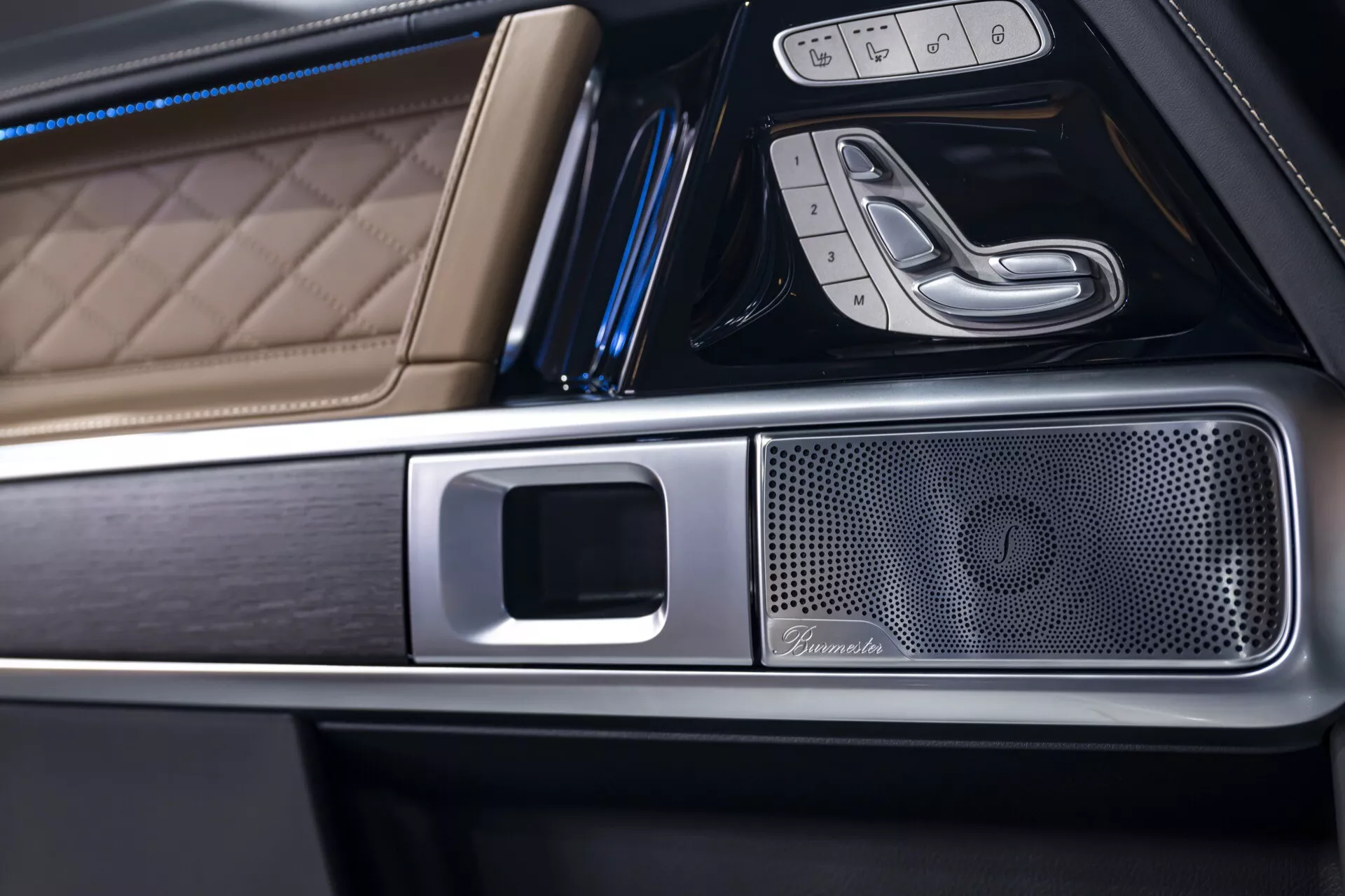 Detailný pohľad na interiér Mercedes-Benz G-Klasse 2024, zdôrazňujúci luxusnú kožu so štvorcovým prešívaním, kovové lemy a prvotriednu mriežku reproduktora na stredovej konzole z uhla