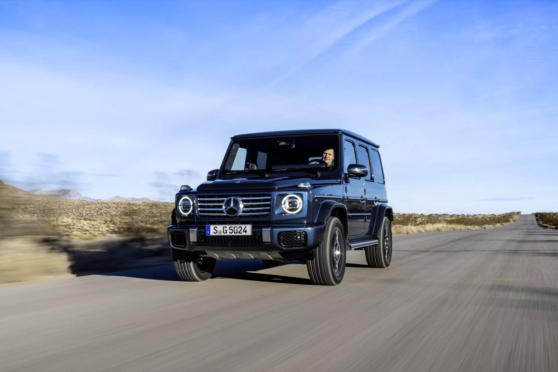 Luxusné SUV Mercedes-Benz triedy G 2024 v pohybe, zachytené z čelného trojštvrtečného uhla, ktoré predvádza LED svetlomety, kruhové denné svetlá, robustný dizajn a pokročilé technologické funkcie na pozadí púštnej cesty