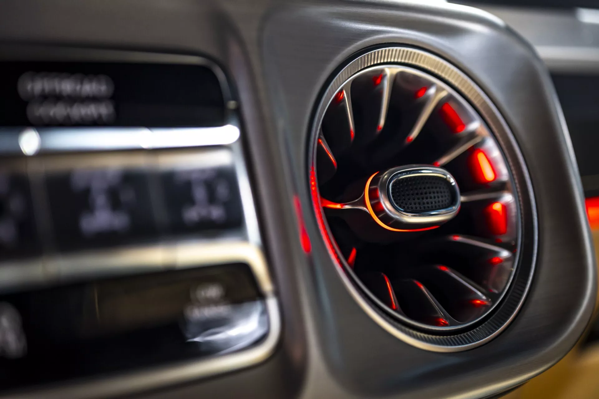 Detailný pohľad na interiér Mercedes-Benz G-Klasse 2024 (465) zobrazujúci vetraciu otvoru s chrómovou povrchovou úpravou a červeným ambientným osvetlením a rozostreným pozadím digitálneho displeja, čo zdôrazňuje luxus a dizajn
