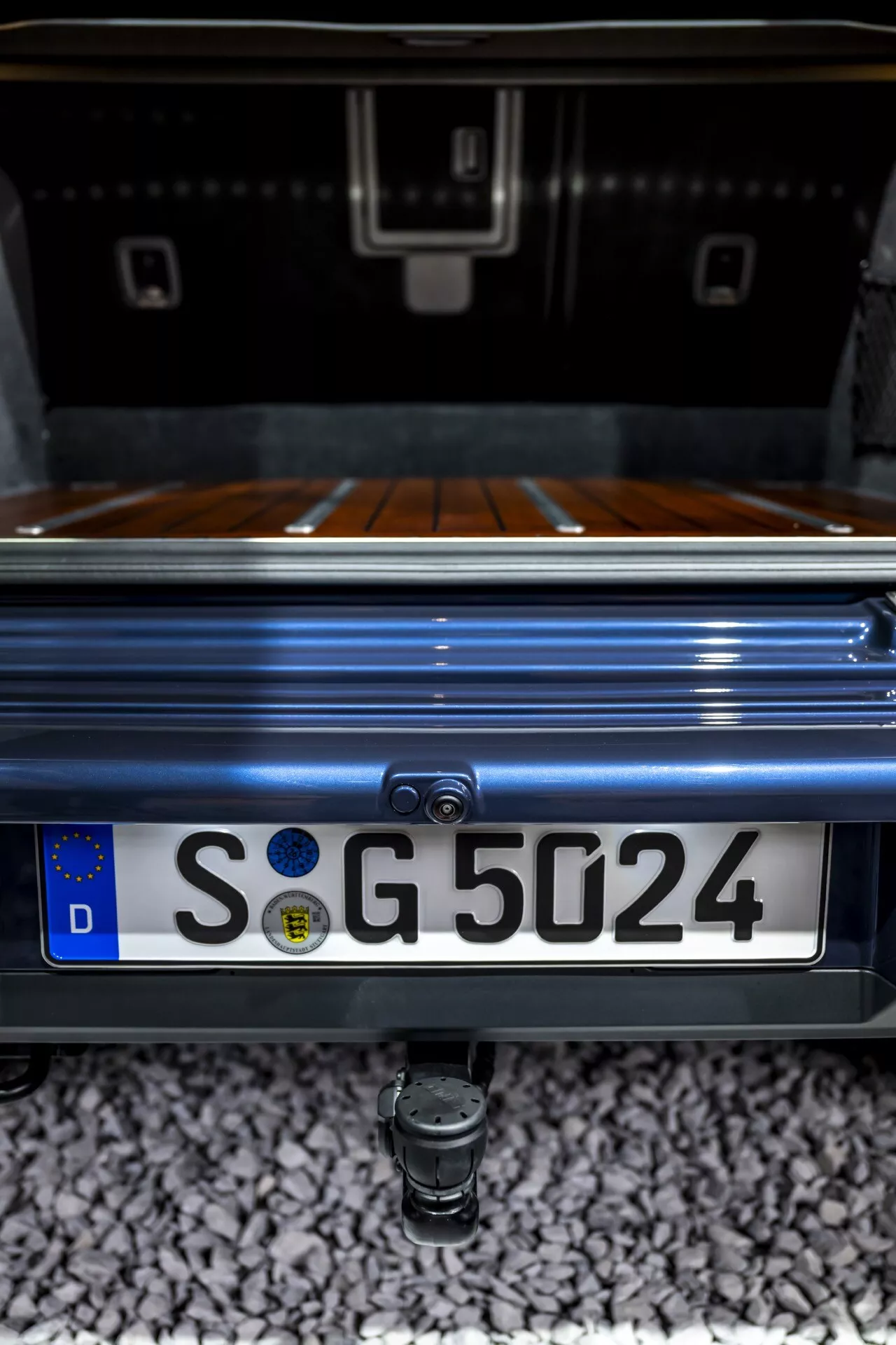 Detailný záber na zadnú časť Mercedes-Benz G-Klasse 2024 so zameraním na priestor batožinového priestoru, luxusné drevené obloženie batožinového priestoru, integrovanú zadnú kameru nad európskou evidenčnou značkou s nemeckým 'D' a ťažné oko nižšie, všetko zachytené z nízkeho, nahor smerujúceho uhla, ktorý zdôrazňuje prémiový metalický lak vozidla a estetiku dizajnu.