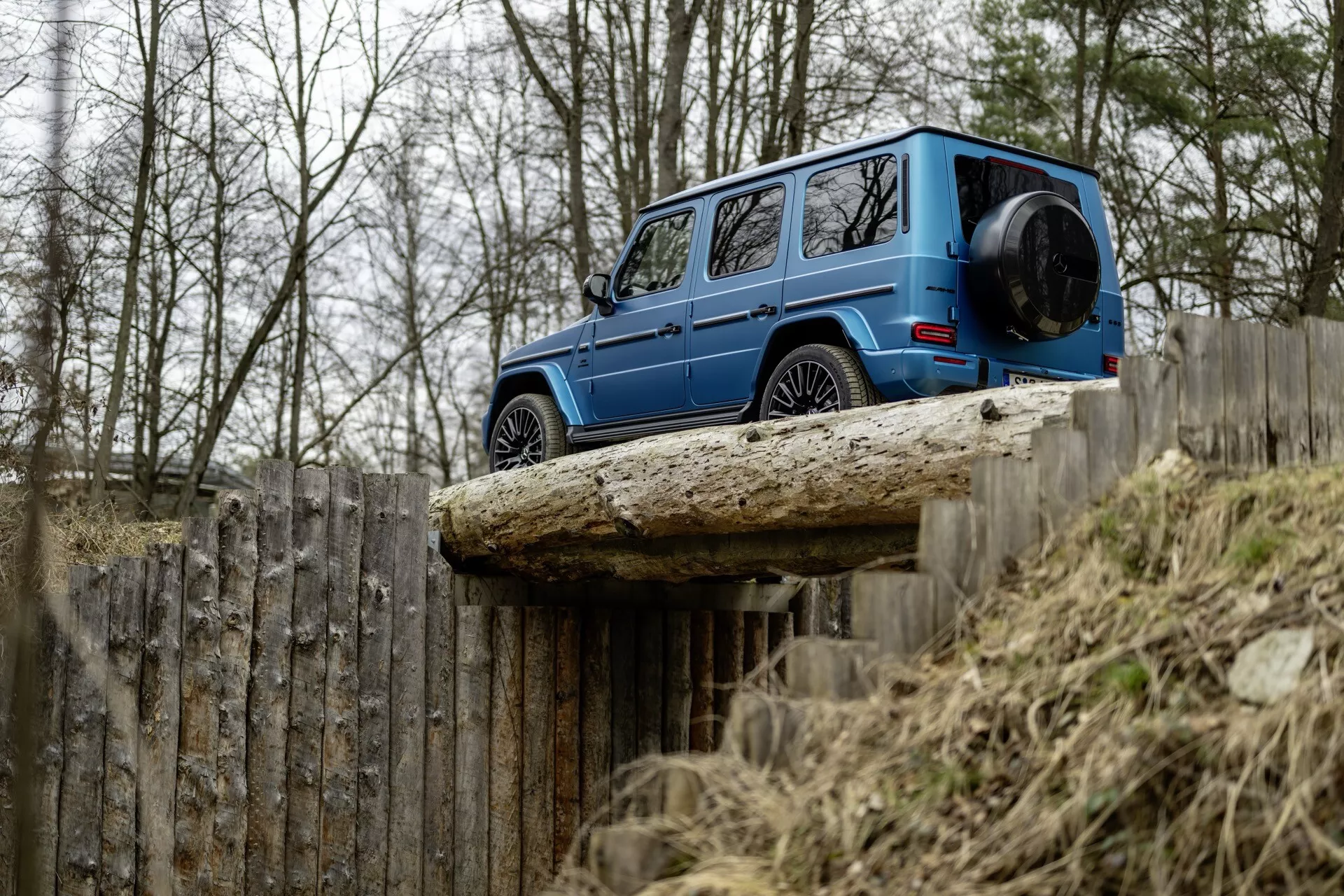 Mercedes-Benz triedy G ročník 2024 v modrej farbe, zobrazený z bočného uhla, ktorý zdôrazňuje jeho škatuľovitý dizajn, odpruženie pre terén a svetlú výšku na zdvihnutej ploche, so zameraním na jeho trvanlivé a luxusné funkcie na pozadí holých stromov a dreveného plotu