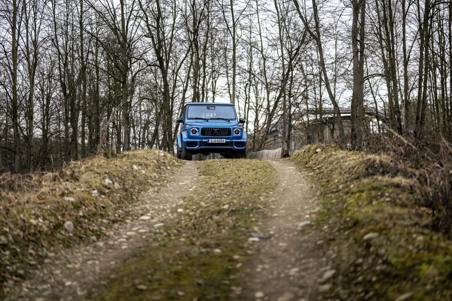 Modré SUV Mercedes-Benz G-Klasse 465 ročník 2024 na lesnej ceste v zalesnenej oblasti predvádzajúce prednú mriežku, LED svetlomety a schopnosti v teréne