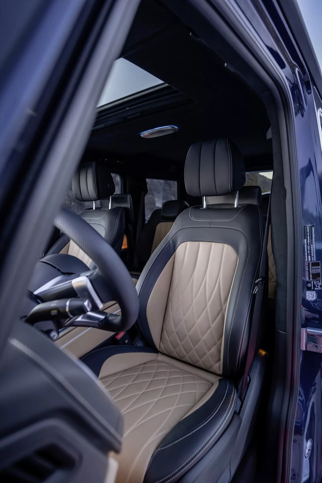Pohled do interiéru Mercedes-Benz Triedy G 465 2024 zo strany vodiča s otvorenými dverami, predstavujúce luxusné čalúnenie a materiály vysokej kvality, ergonomický volant a sofistikované dizajnové prvky